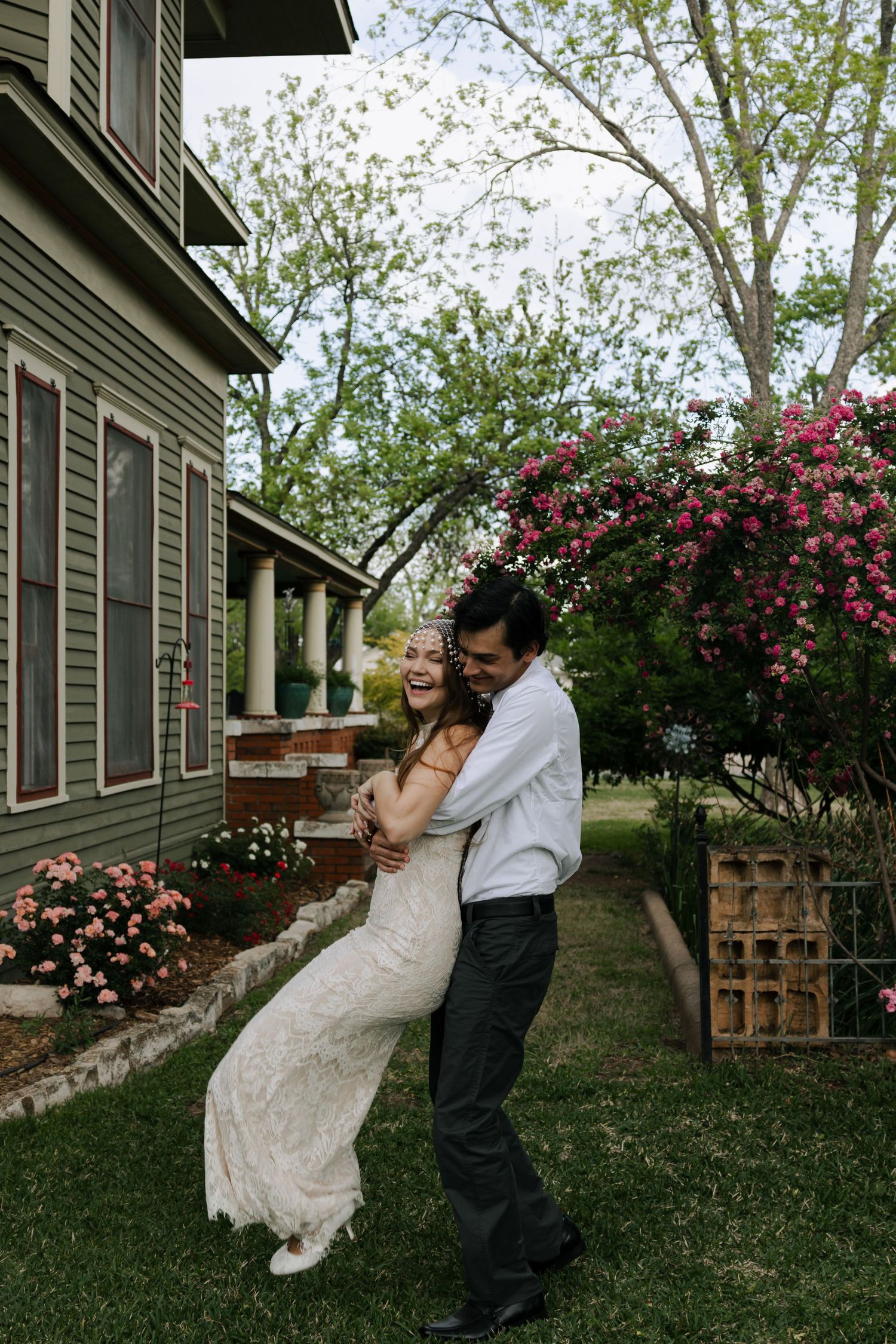 Happy bride and groom embracing in a lush garden with pink flowering bushes outside a green house.