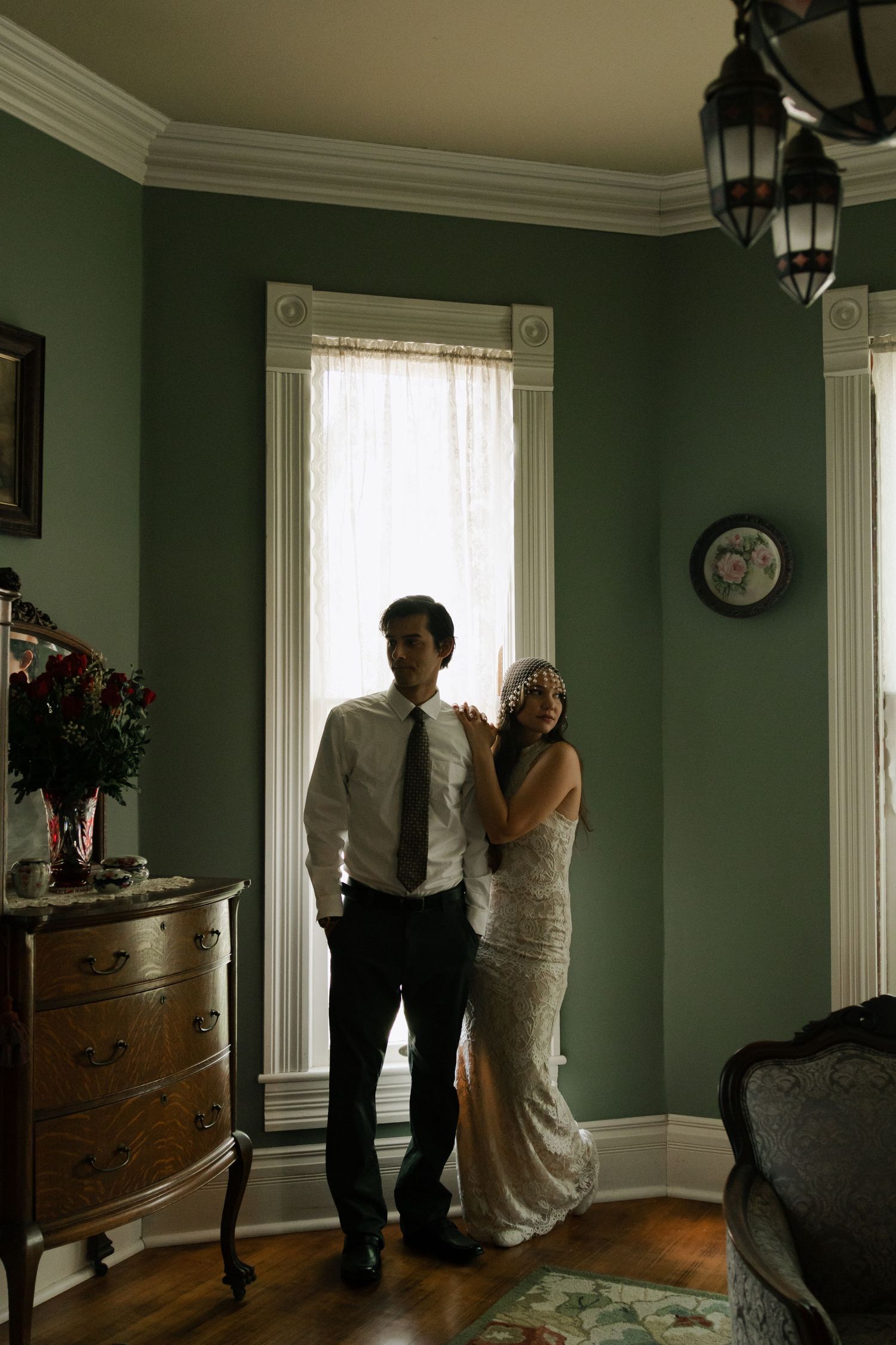 Bride and groom stand together in elegant green-walled room with vintage dresser and chandelier in moody wedding portrait.