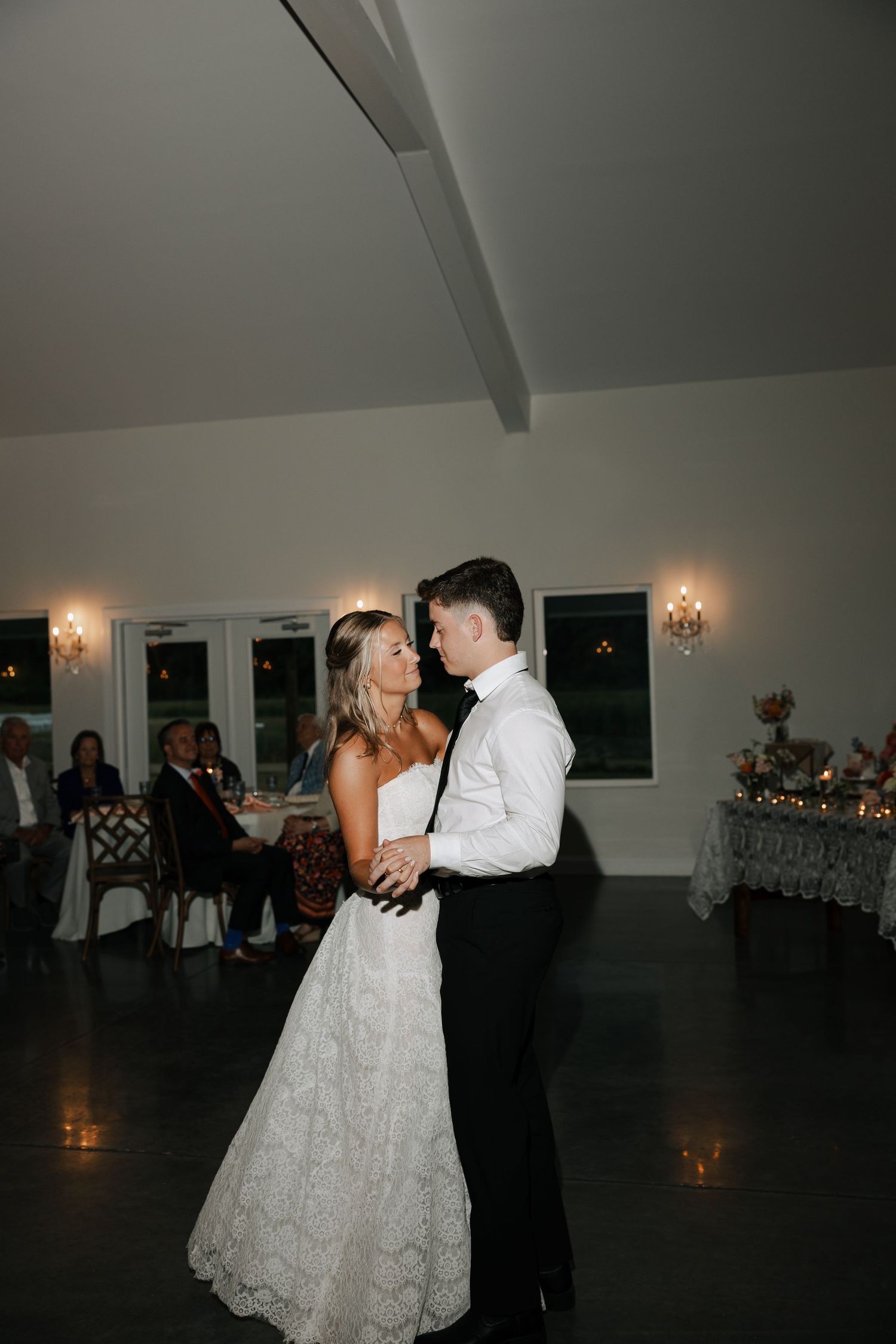 Bride and groom share their first dance in an elegant wedding reception venue with warm candlelight and guests seated nearby.
