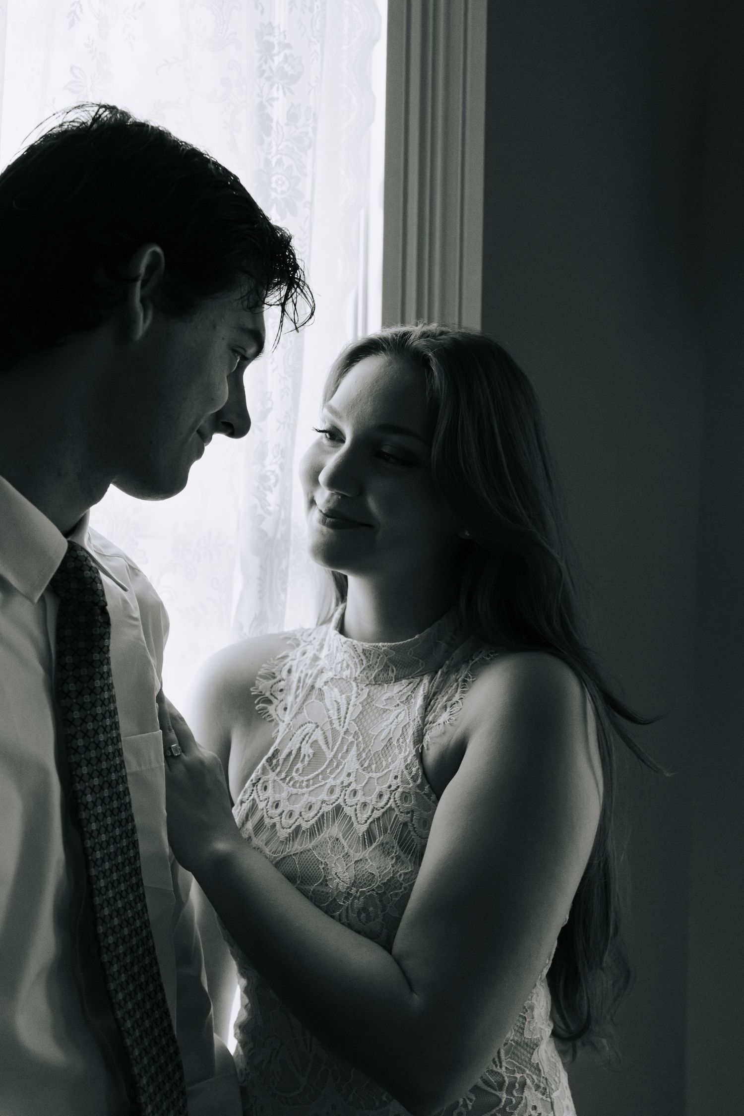 Black and white photo of a bride and groom sharing an intimate moment by a window.