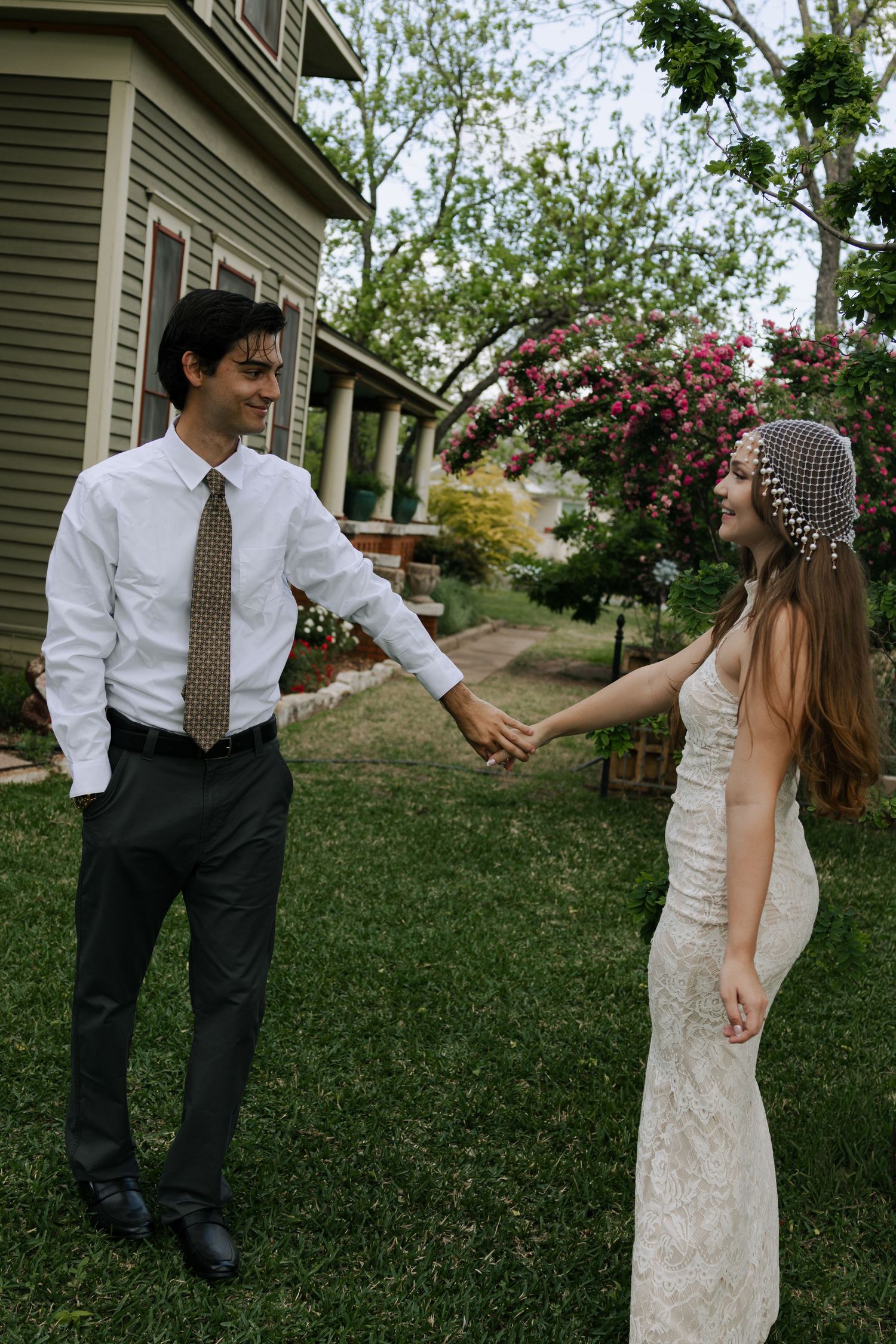 Bride and groom hold hands in a garden, bride in lace dress with pearl headpiece, groom in white shirt and tie.