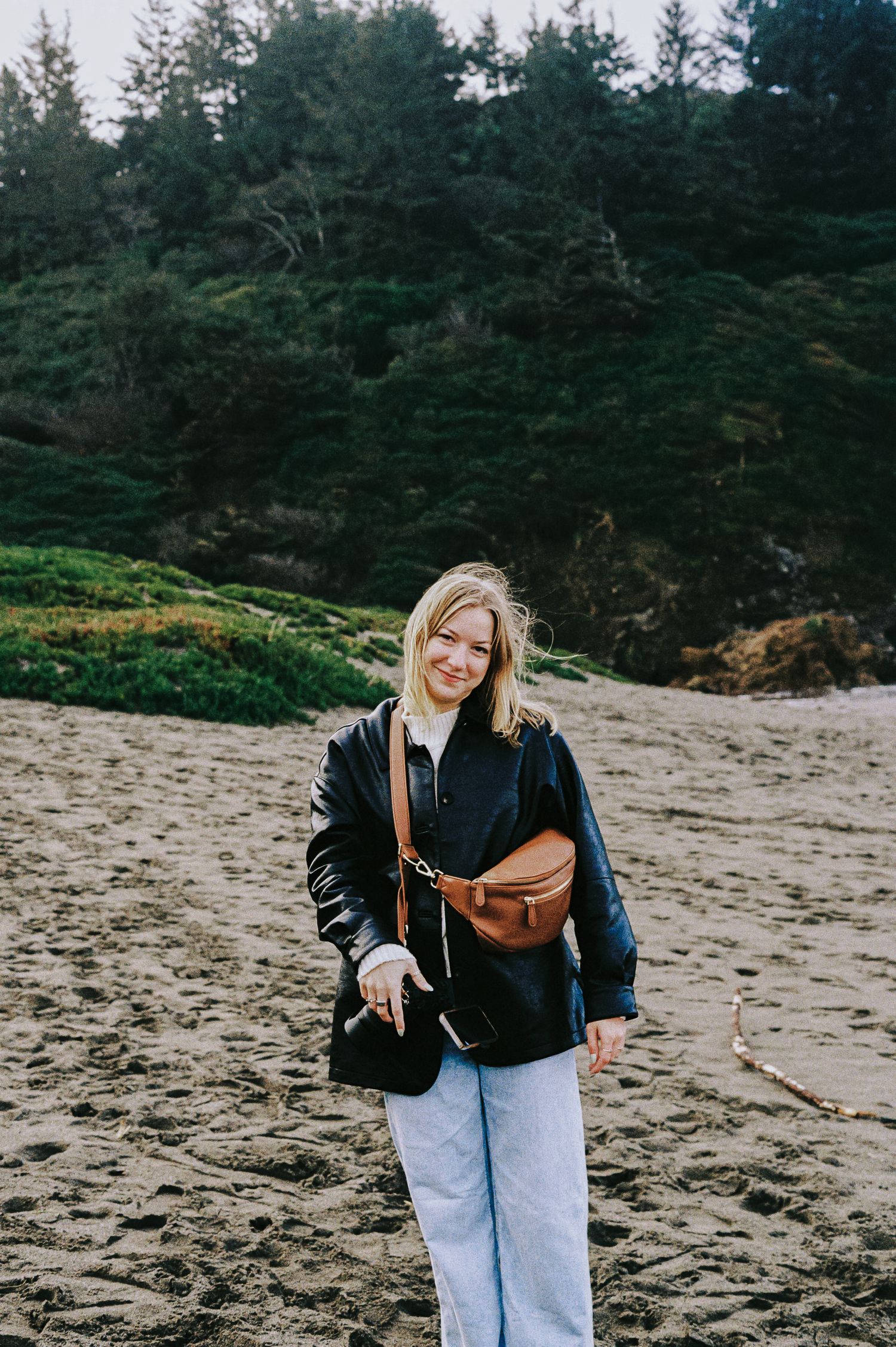 Smiling blonde woman in black leather jacket with brown belt bag on sandy beach.