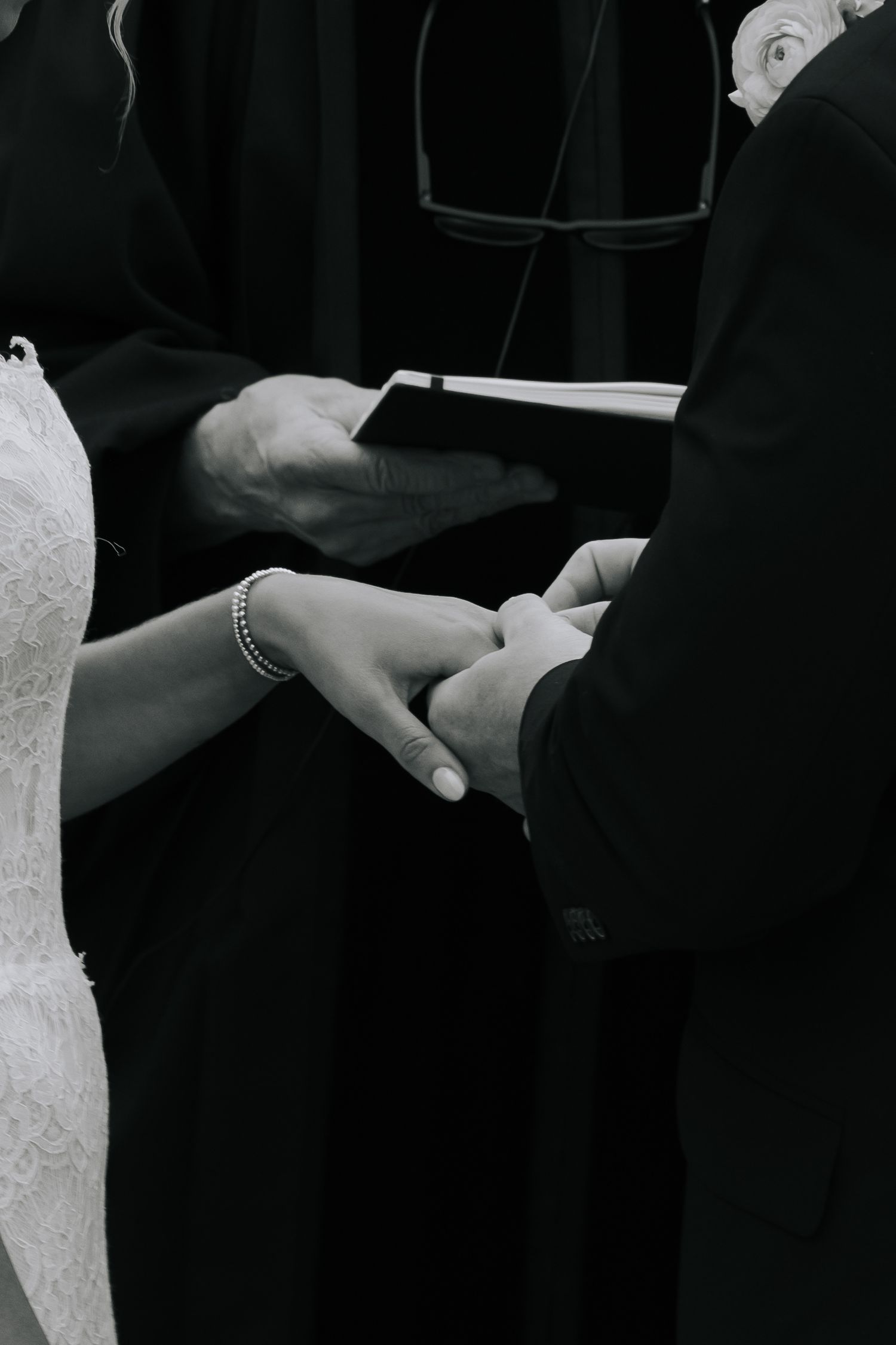 Black and white photo of bride and groom holding hands during wedding ring exchange ceremony.