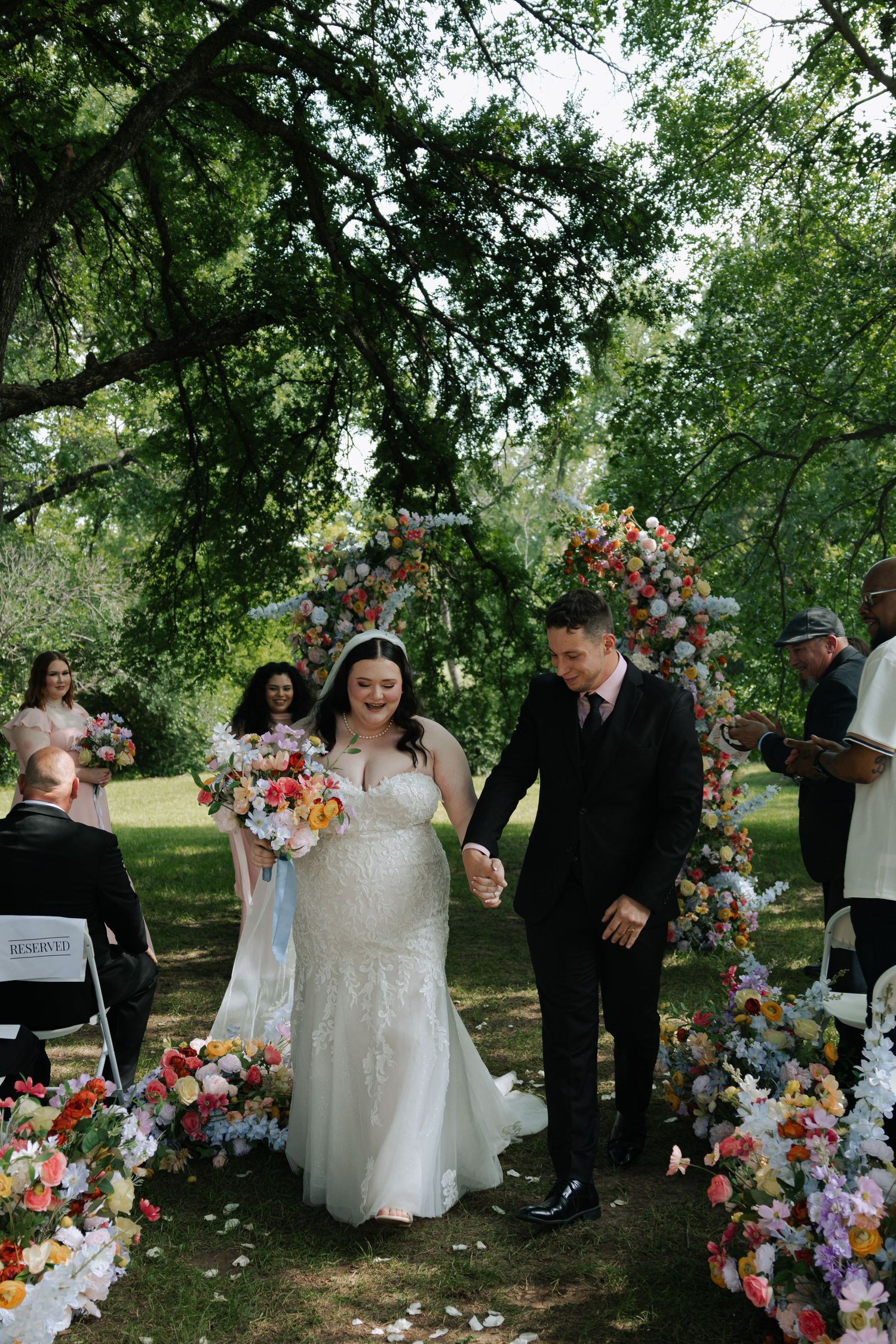 Bride and groom walking through a colorful floral arch after their outdoor garden wedding ceremony.
