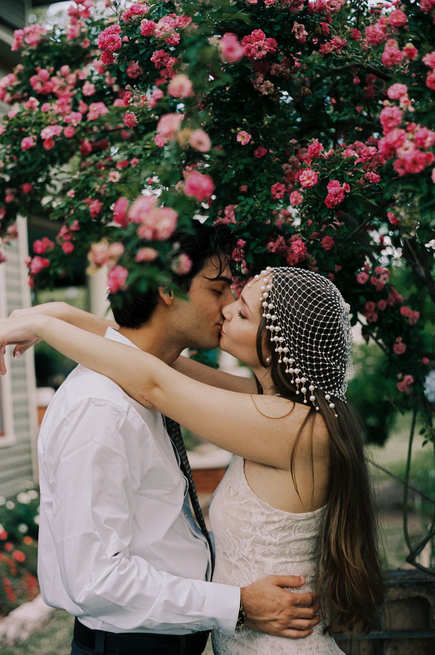 Bride and groom share a romantic kiss under a blooming pink rose bush, bride wearing an elegant beaded veil headpiece.