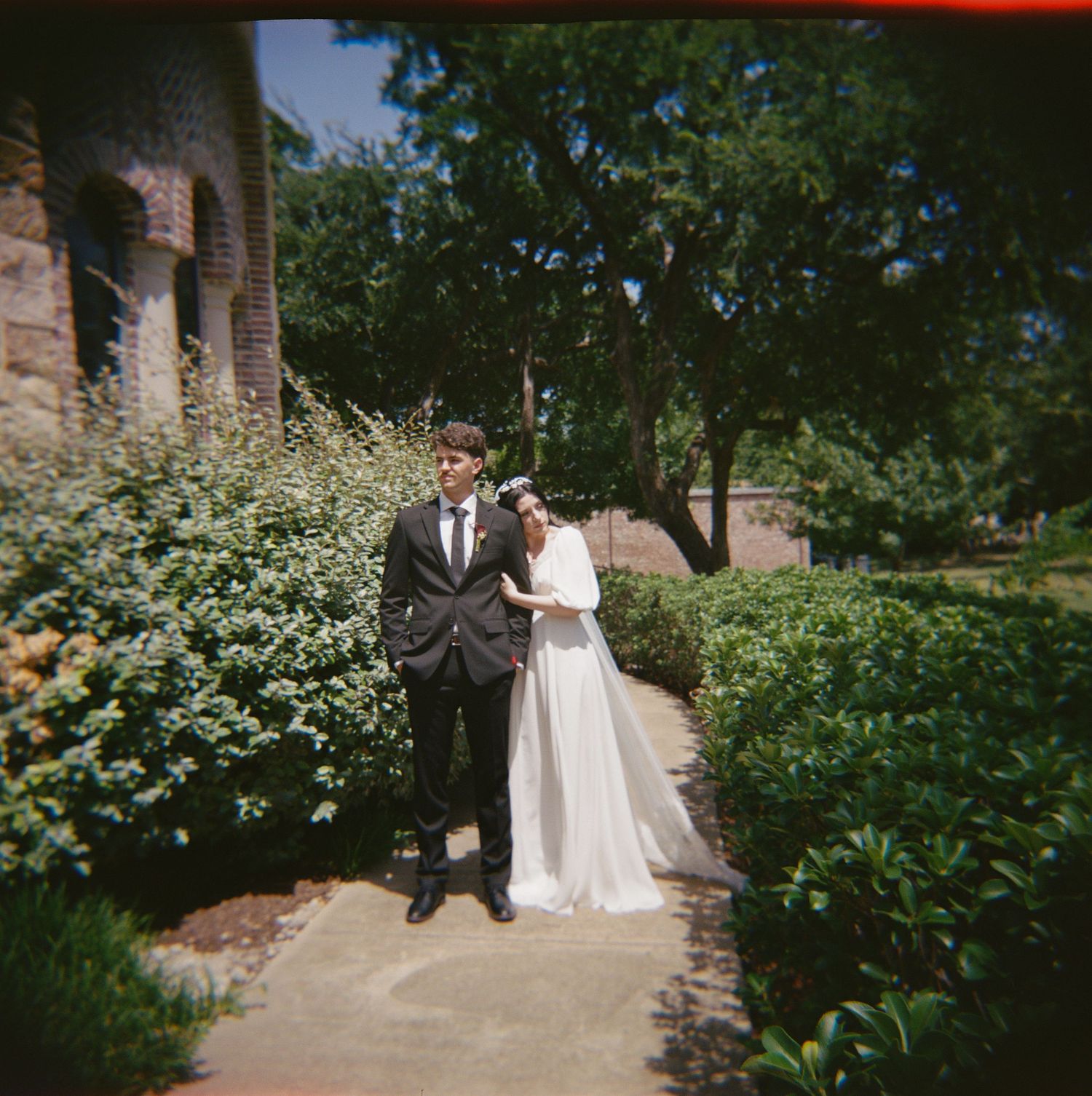 A bride and groom pose together in a lush garden pathway beside a brick building on their wedding day.