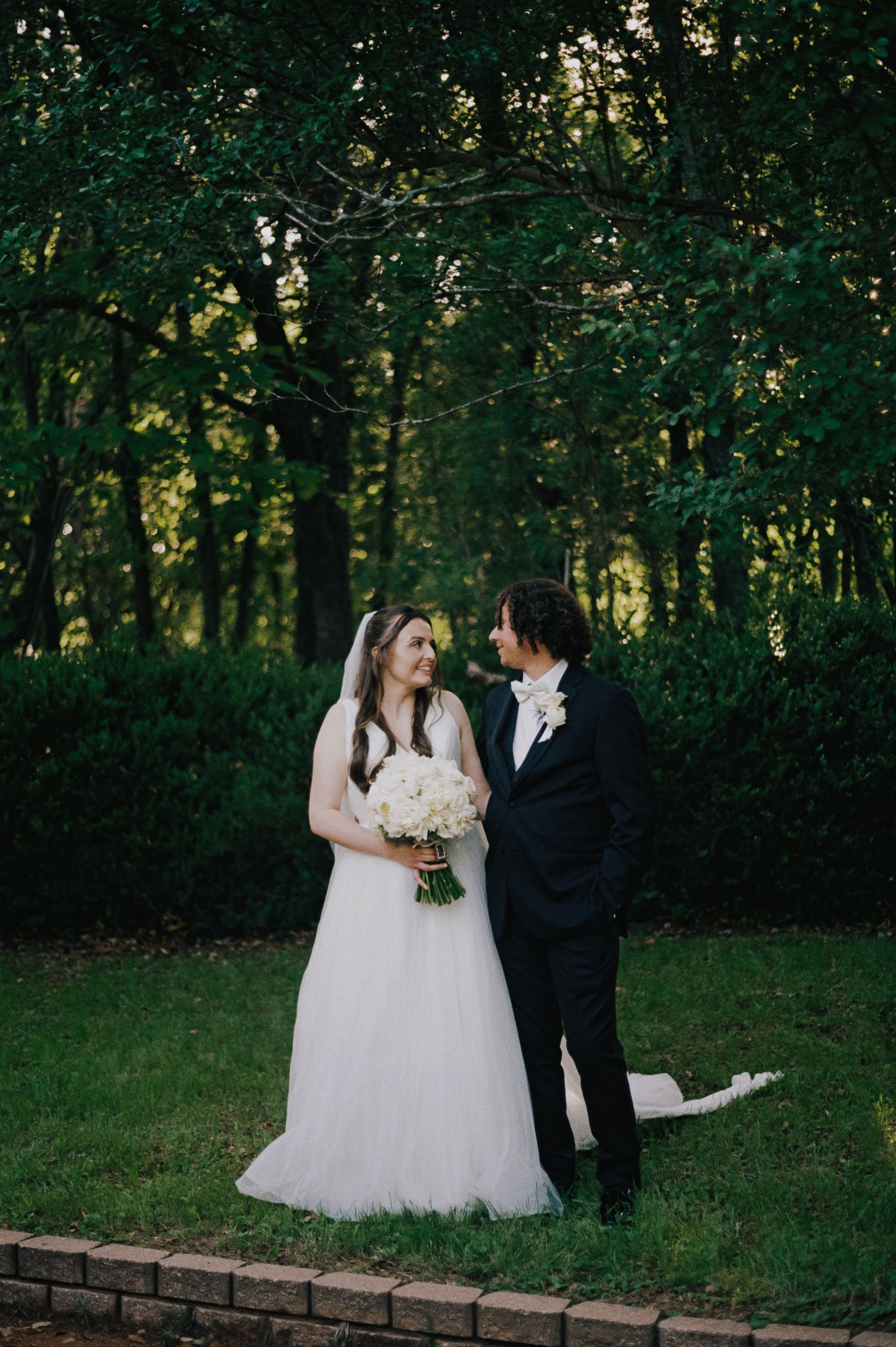 Bride in white gown holding bouquet stands with groom in black tuxedo outdoors, sharing a romantic moment before lush green trees.