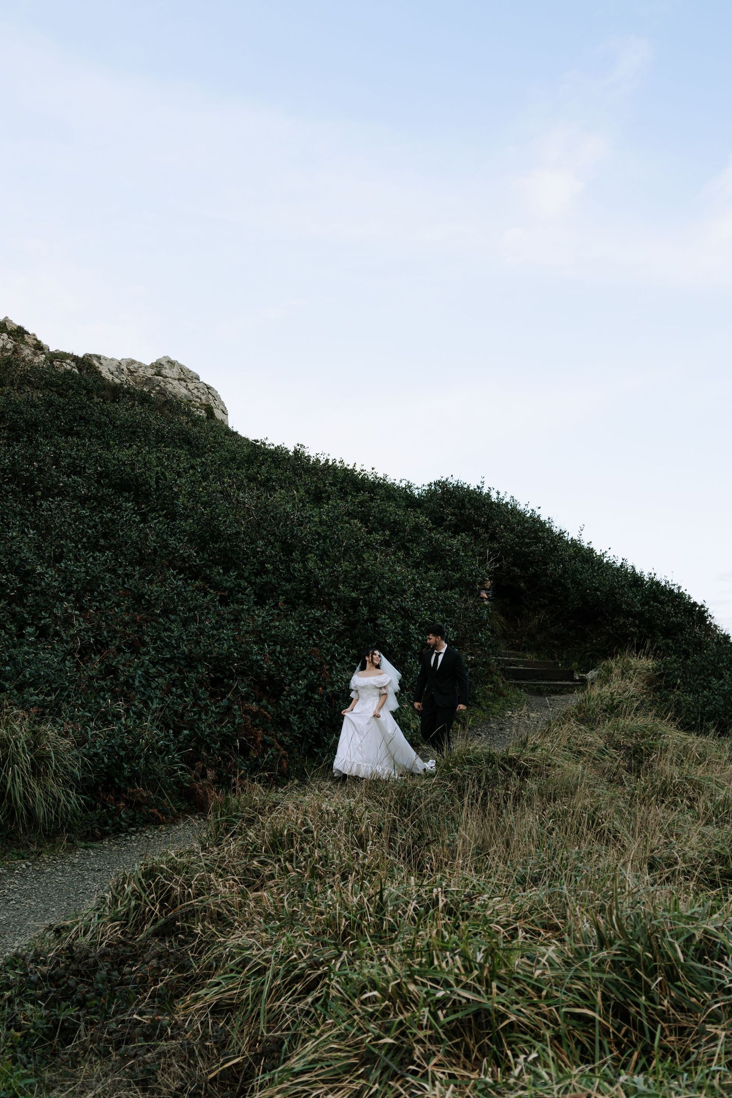 Bride and groom walk hand-in-hand along a lush green hillside path on their wedding day.