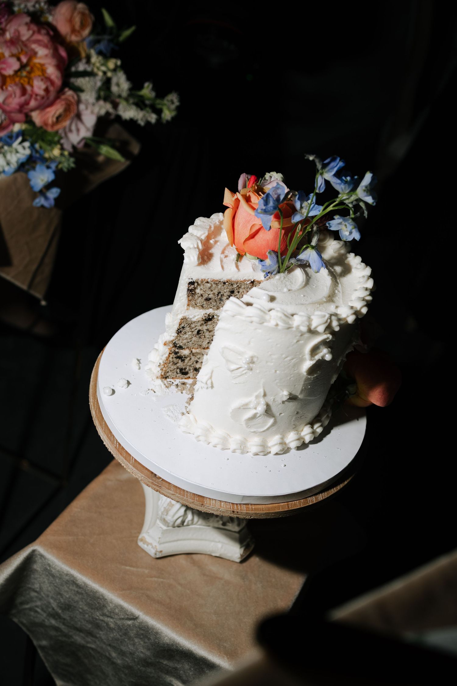 Elegant single-tier white wedding cake slice topped with peach rose and blue flowers on a decorative stand.