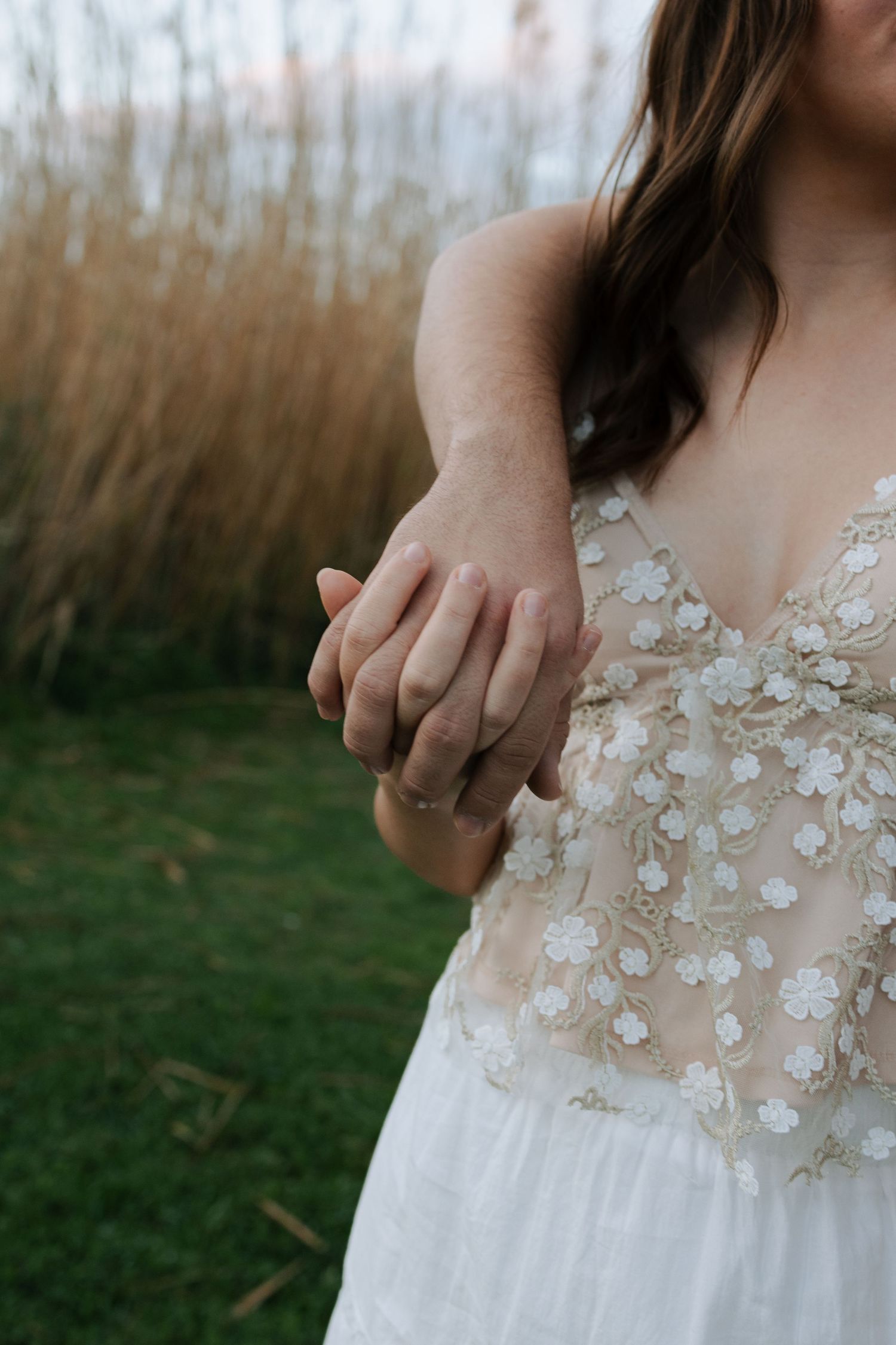 Close-up of engaged couple holding hands, woman wearing floral embroidered wedding dress outdoors.
