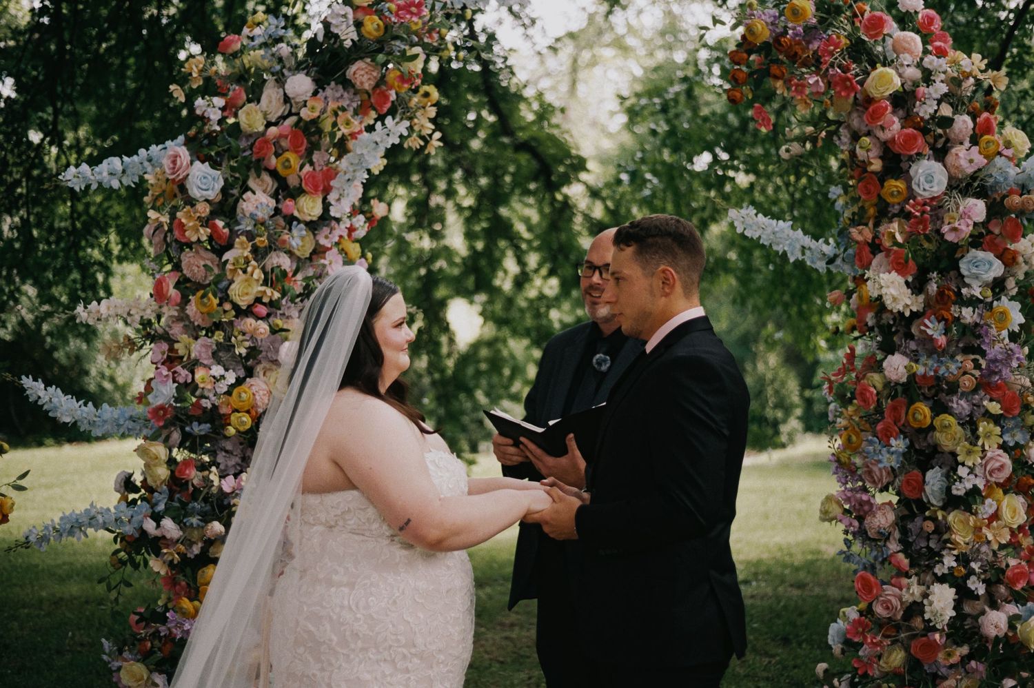 Bride and groom exchange vows under a colorful floral arch outdoors during a romantic garden wedding ceremony.