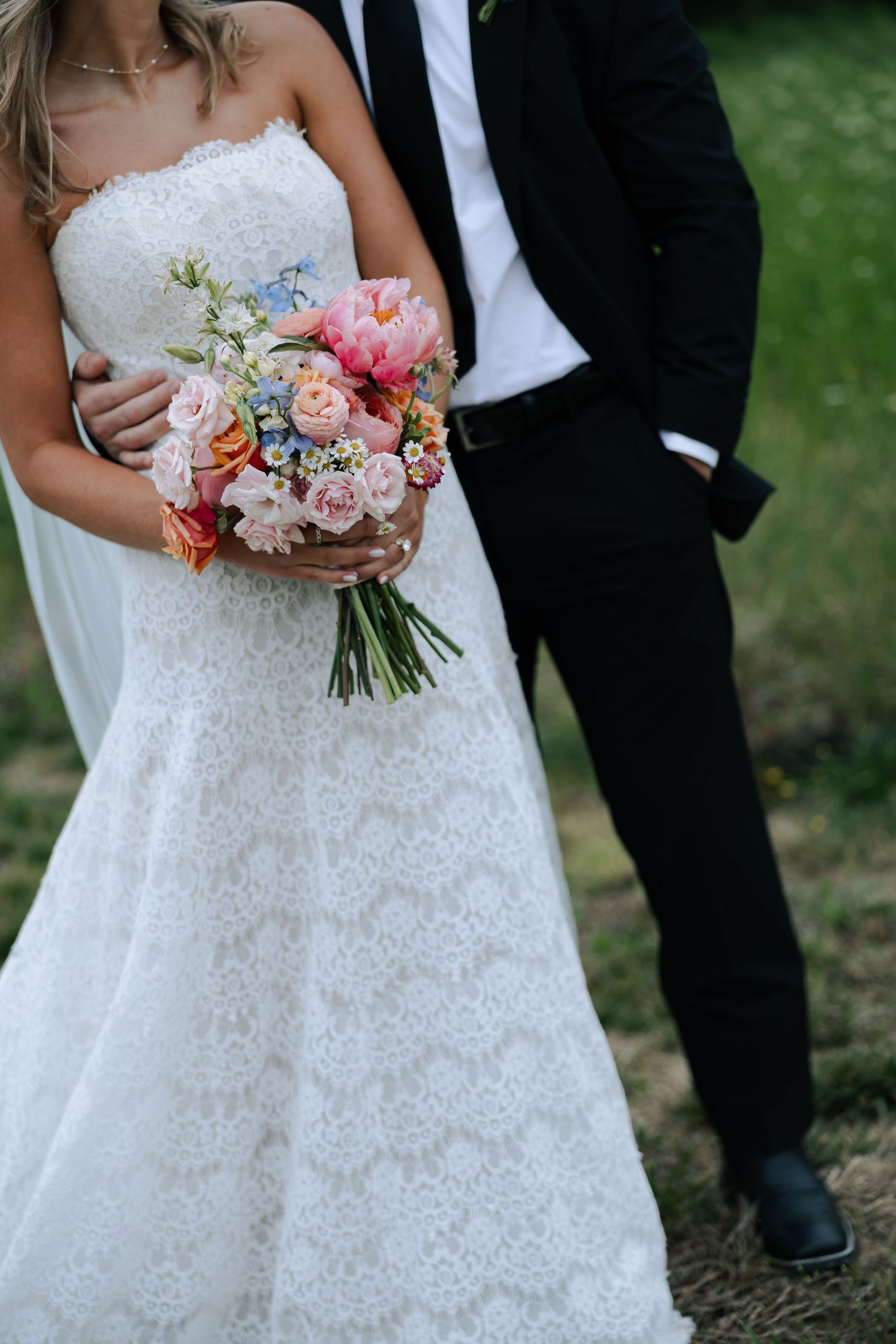 Bride in strapless lace gown holds colorful pink and orange bouquet while groom embraces her.