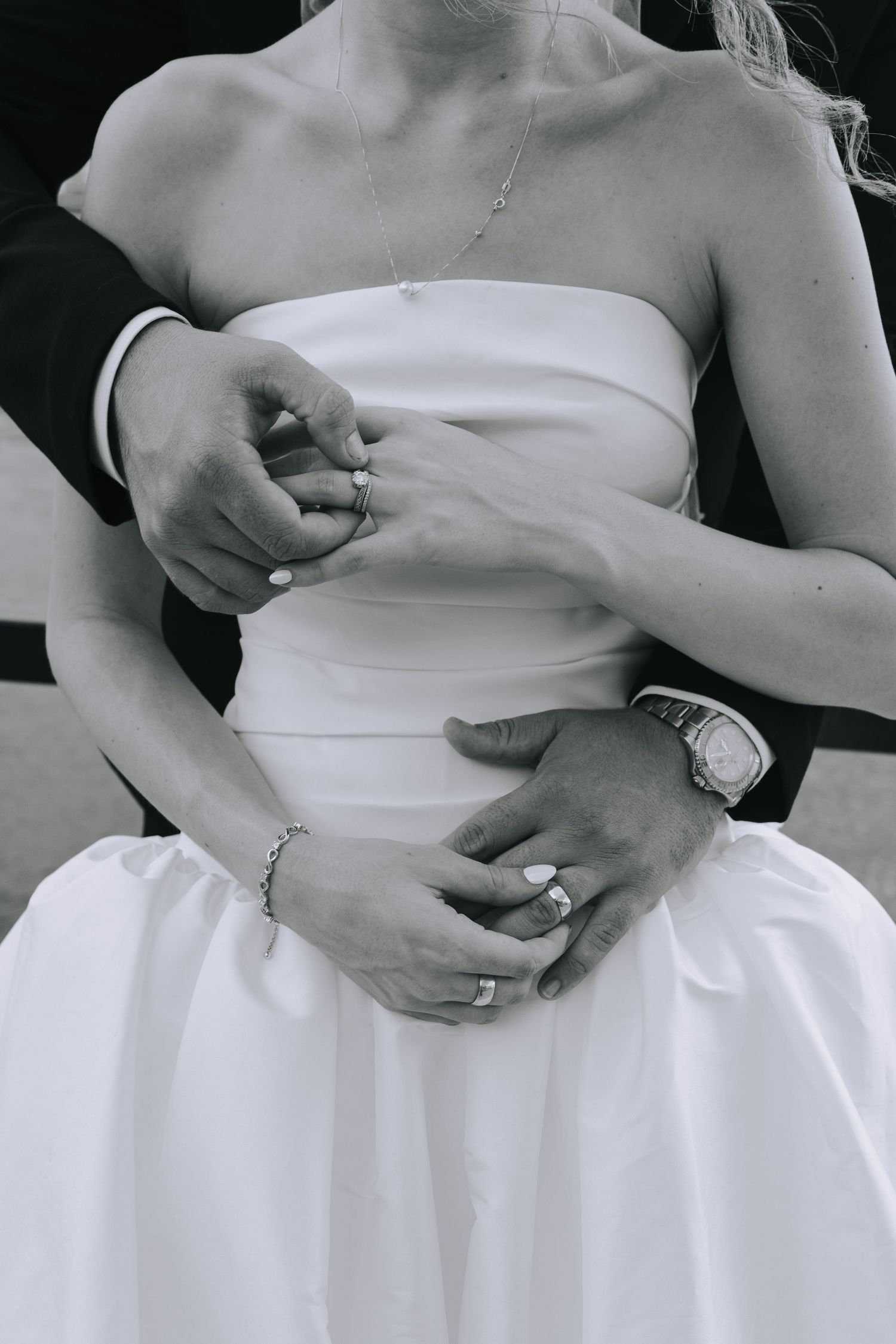 Black and white close-up of bride and groom's hands showing wedding rings, bride in strapless white gown.