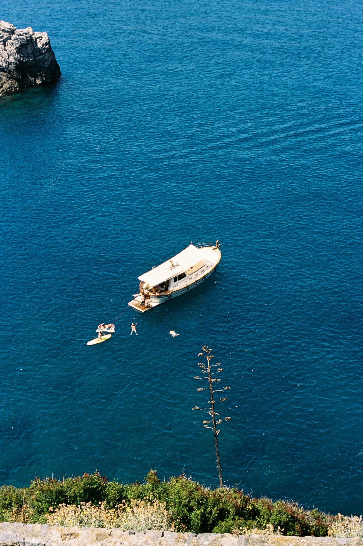 Aerial view of a white motorboat anchored in vivid blue Mediterranean sea waters near rocky cliffs with swimmers nearby.