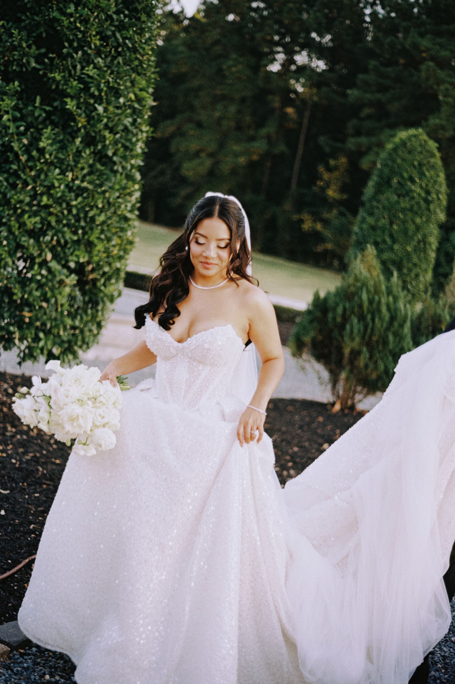 Bride in blush sparkle strapless ball gown holding white bouquet outdoors among lush green hedges.