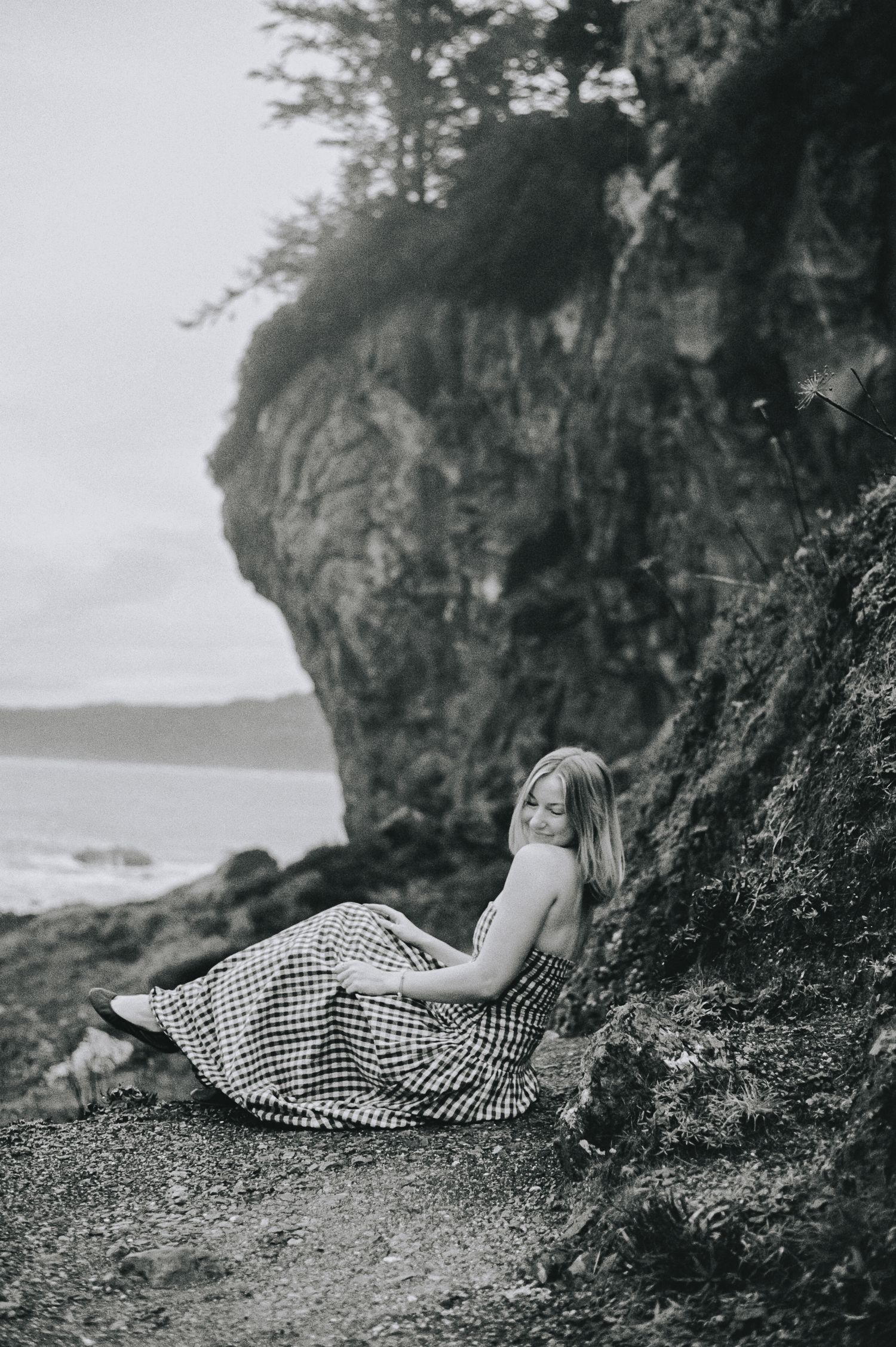 Woman in plaid dress sitting against rocky coastal cliff in black and white photography.