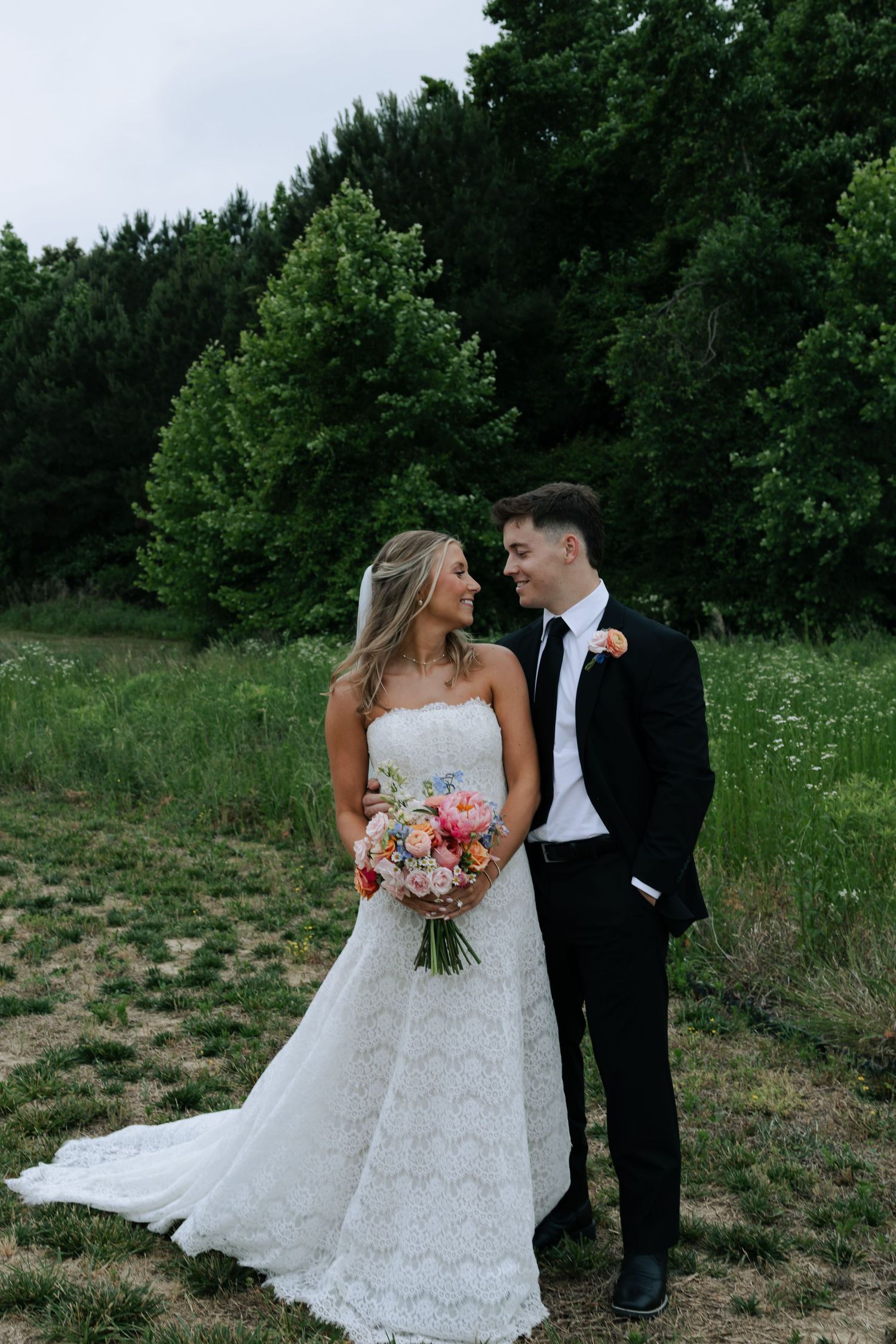 Bride and groom share a romantic moment outdoors, surrounded by lush green trees, as she holds a colorful wedding bouquet.