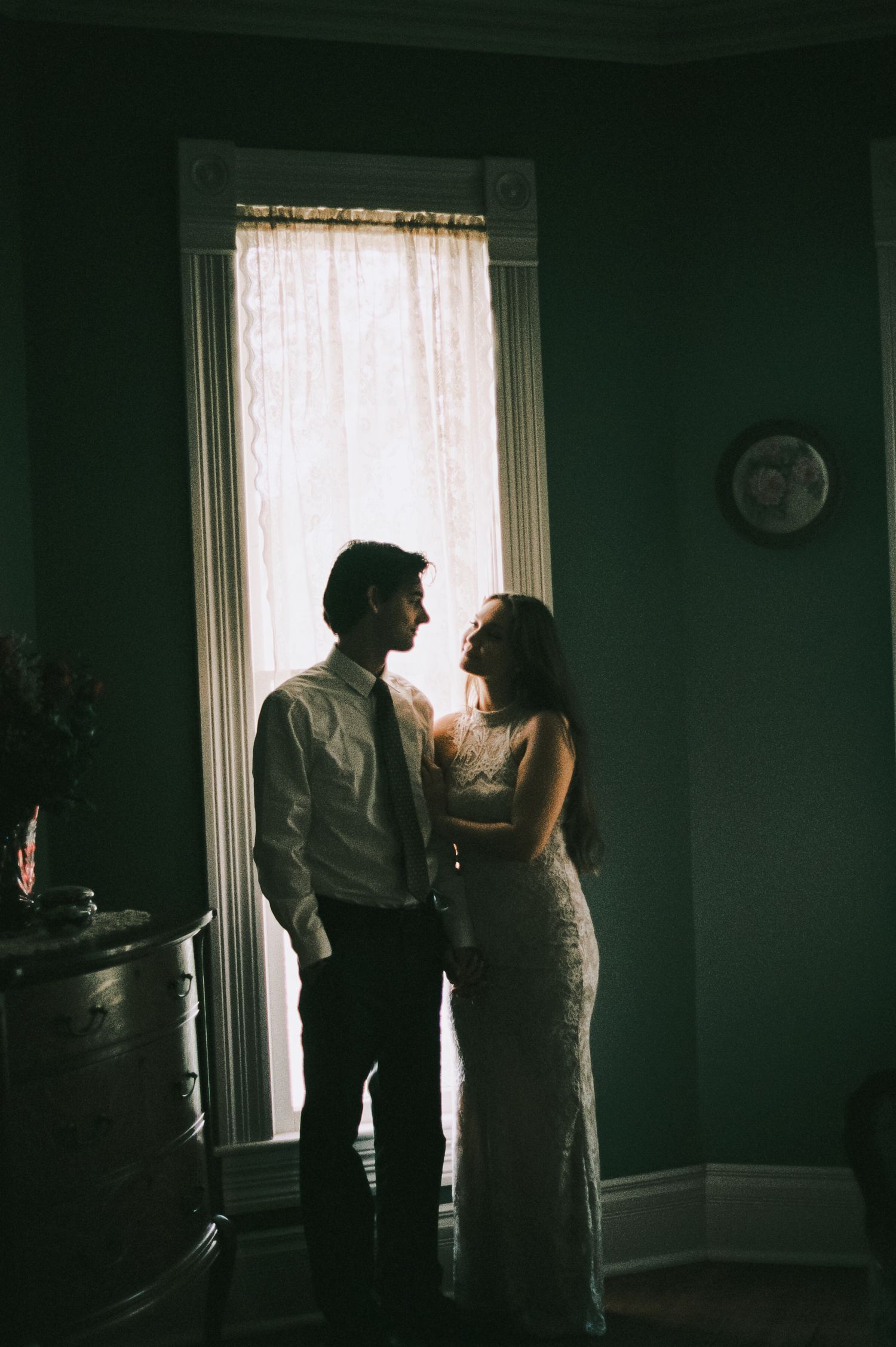 Romantic couple silhouetted against a bright window in a dimly lit room, sharing an intimate moment in wedding attire.