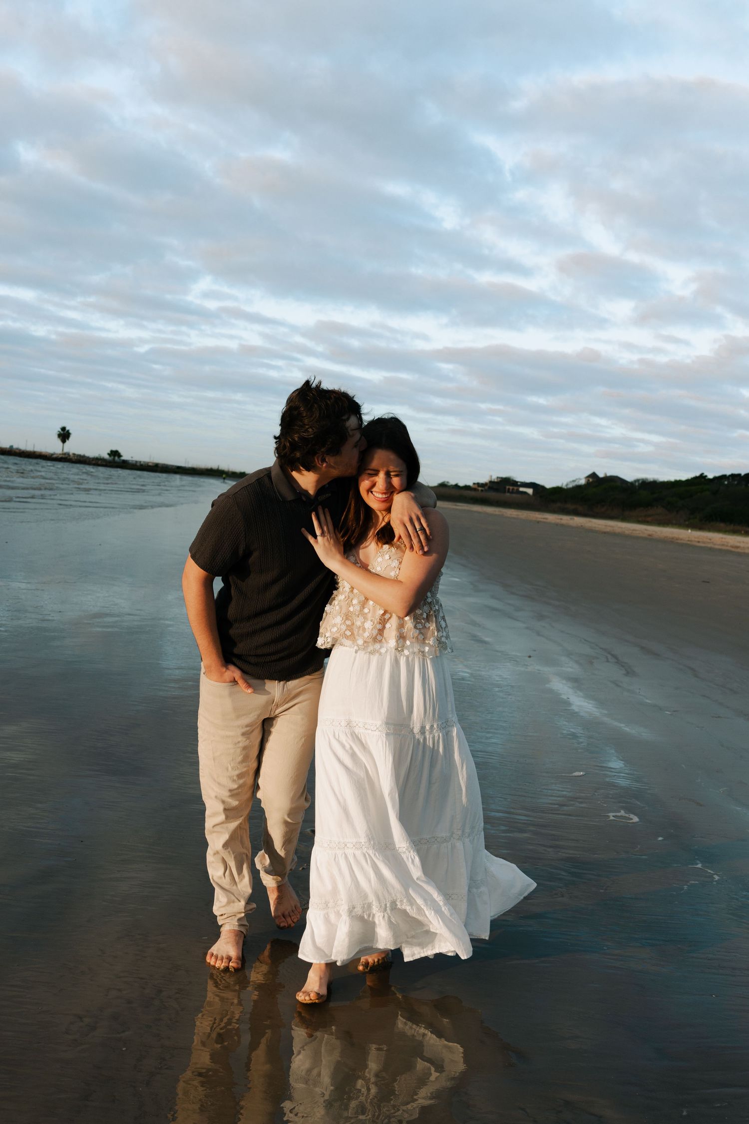 Romantic couple embracing on a wet sandy beach, woman in white dress smiling as man kisses her cheek.