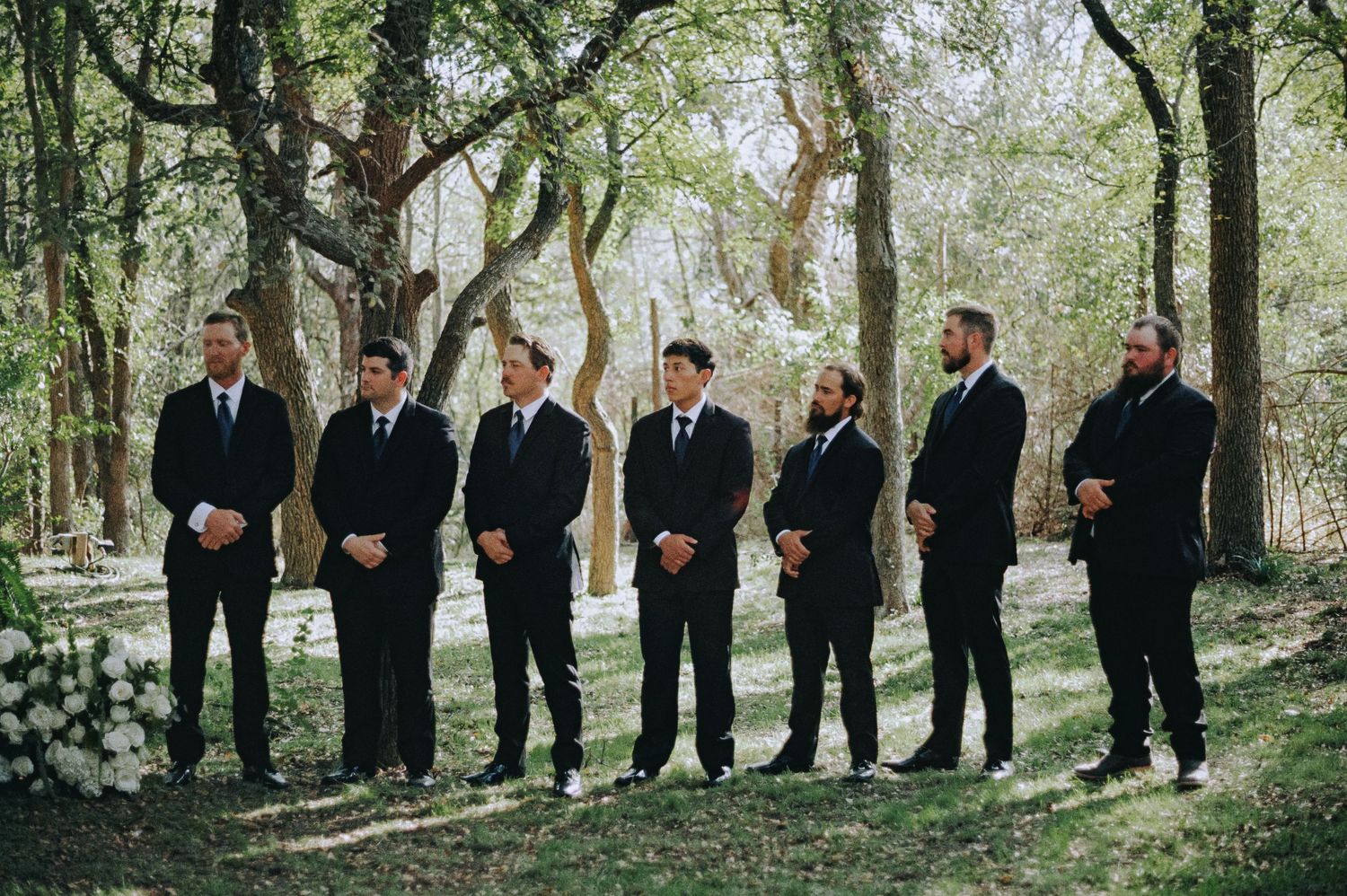 Groomsmen in matching black suits stand outdoors in a wooded wedding ceremony setting with white floral arrangements.