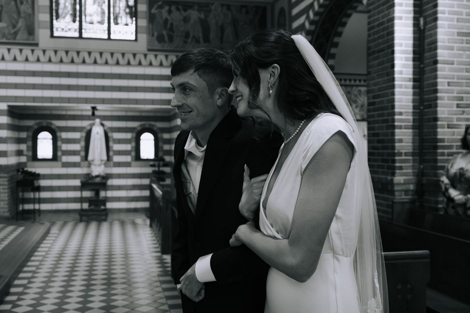 Black and white photo of a smiling groom and bride in veil sharing a moment inside a church.