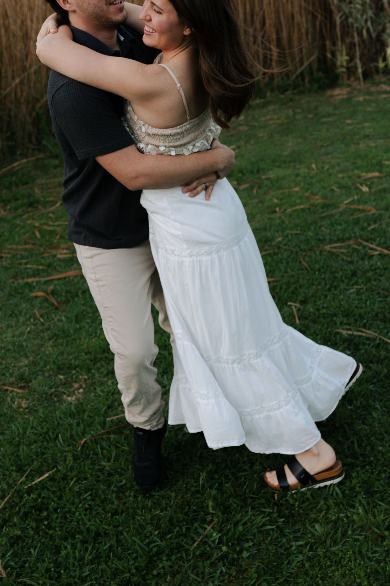 Couple embracing and laughing together in front of tall reeds during outdoor engagement photo session.
