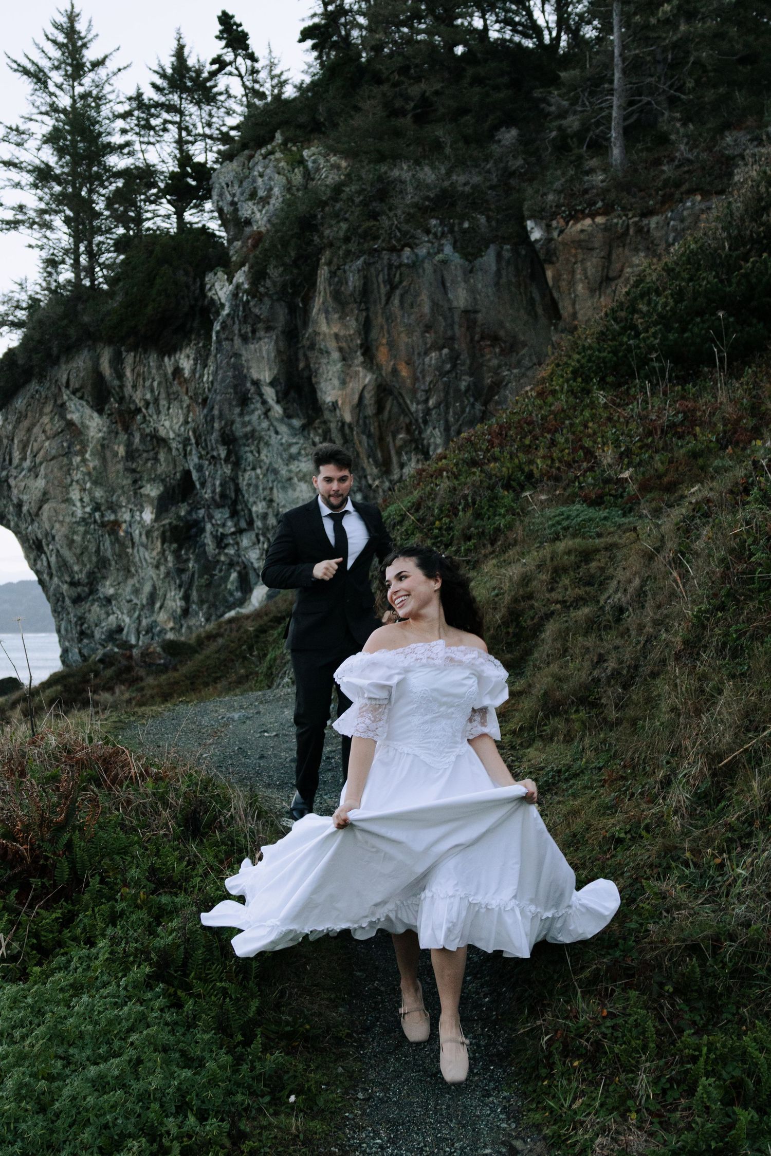 Bride in white dress holds skirt while walking coastal path with groom in black suit behind rocky cliffs.