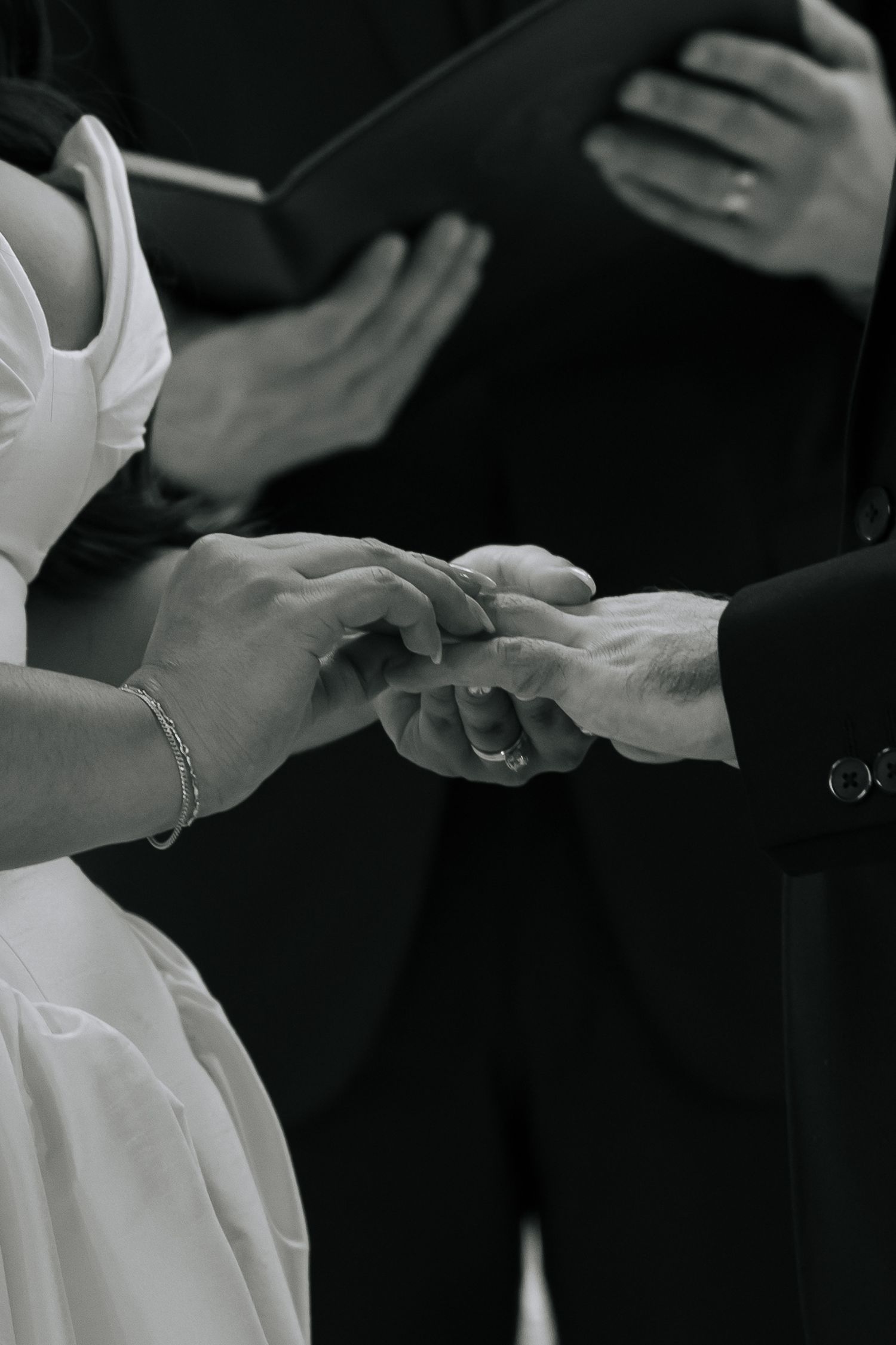 Black and white photo of a couple exchanging wedding rings during a marriage ceremony with officiant in background.
