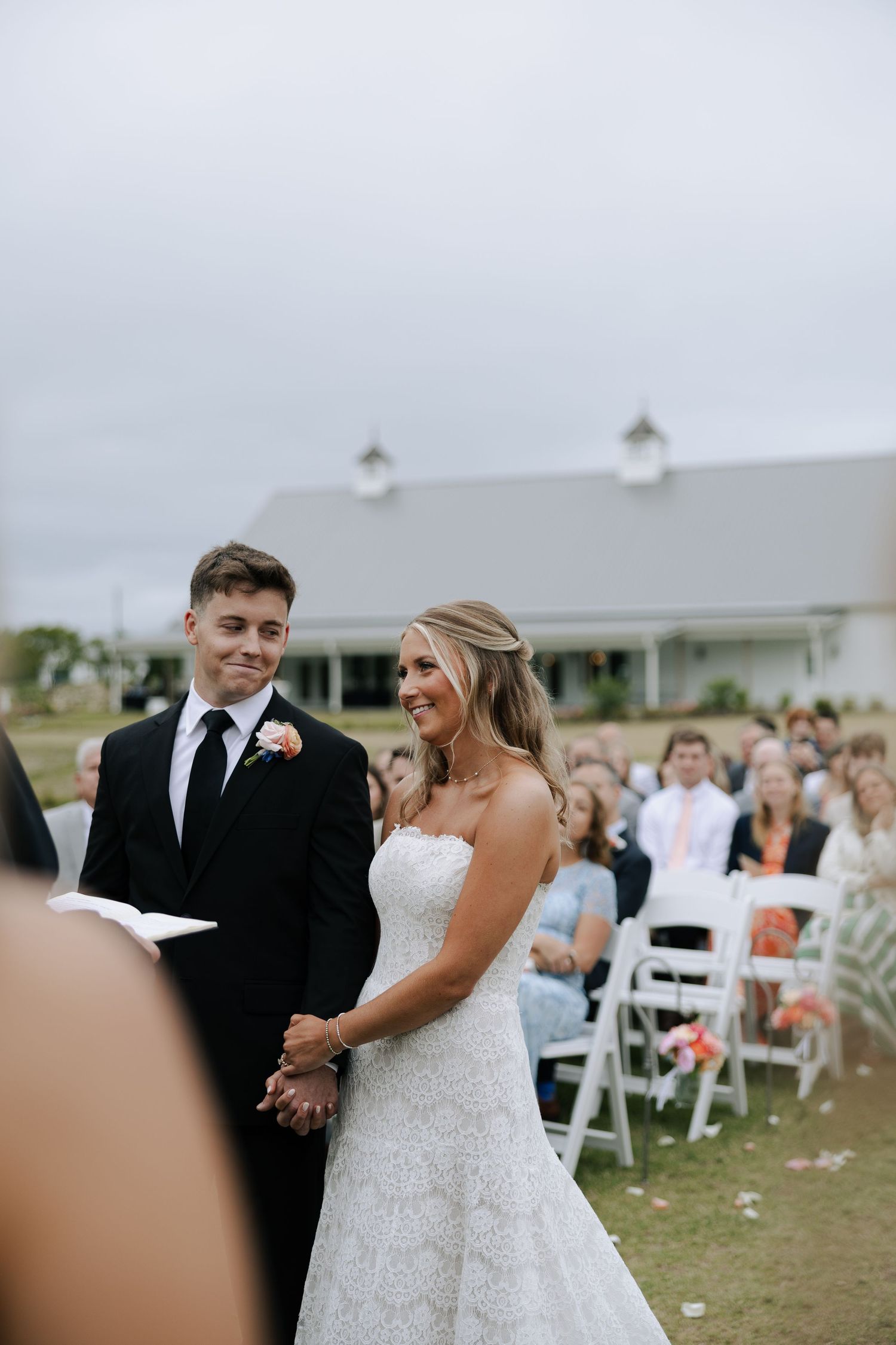 Bride and groom smiling during outdoor wedding ceremony with seated guests in background.