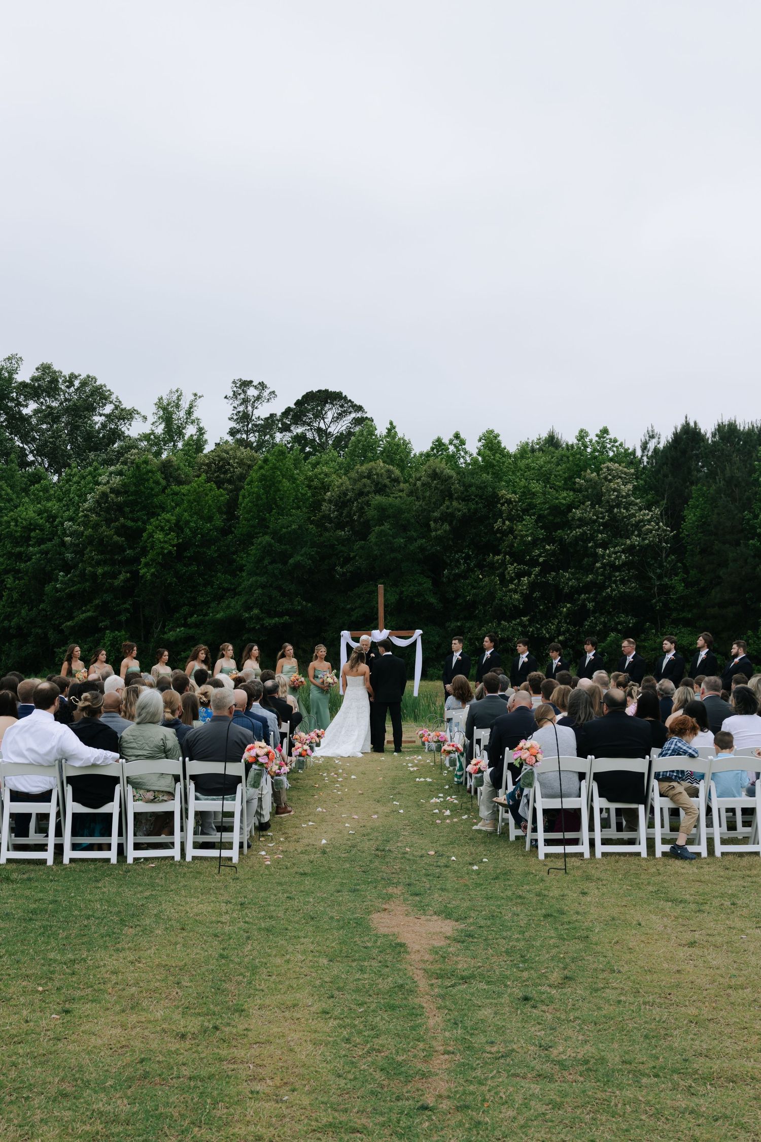 Outdoor wedding ceremony on a green lawn with guests seated in white chairs facing the altar amid lush trees.