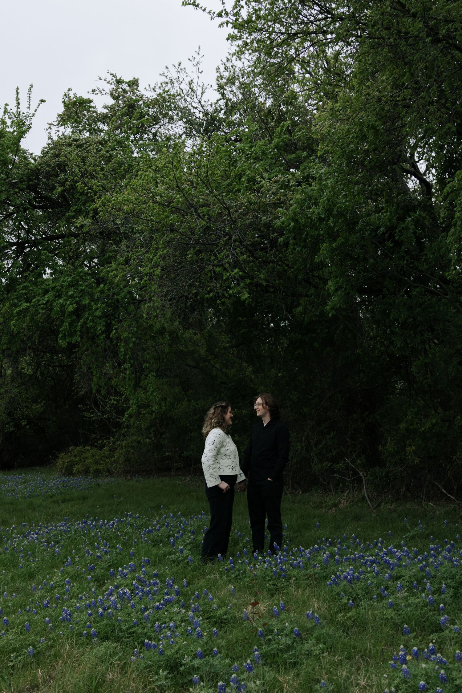 Couple holding hands in a field of bluebonnets surrounded by lush green trees at dusk.