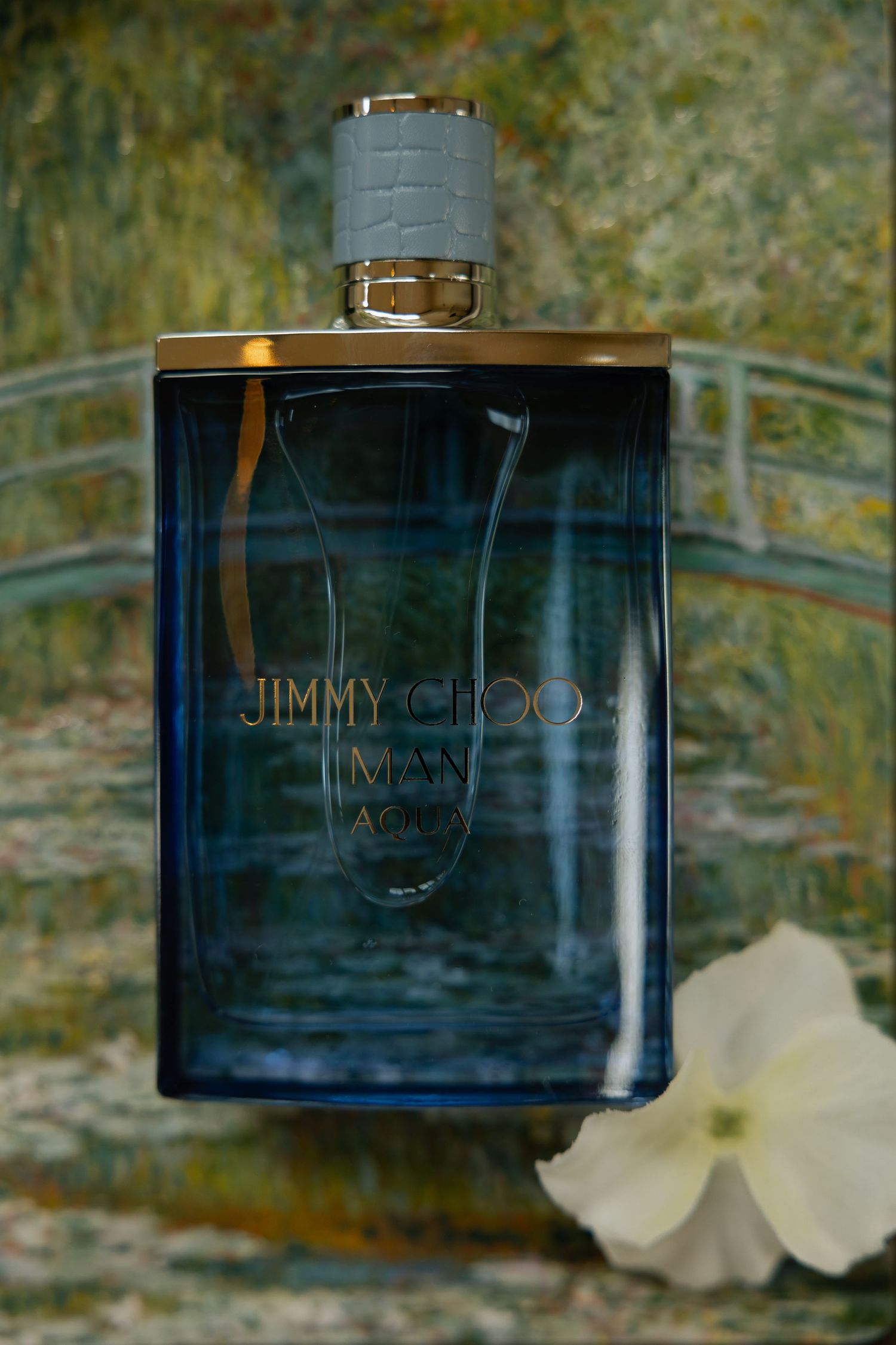 Jimmy Choo Man Aqua cologne in a deep blue bottle with gold accents, displayed against a Monet-inspired background.