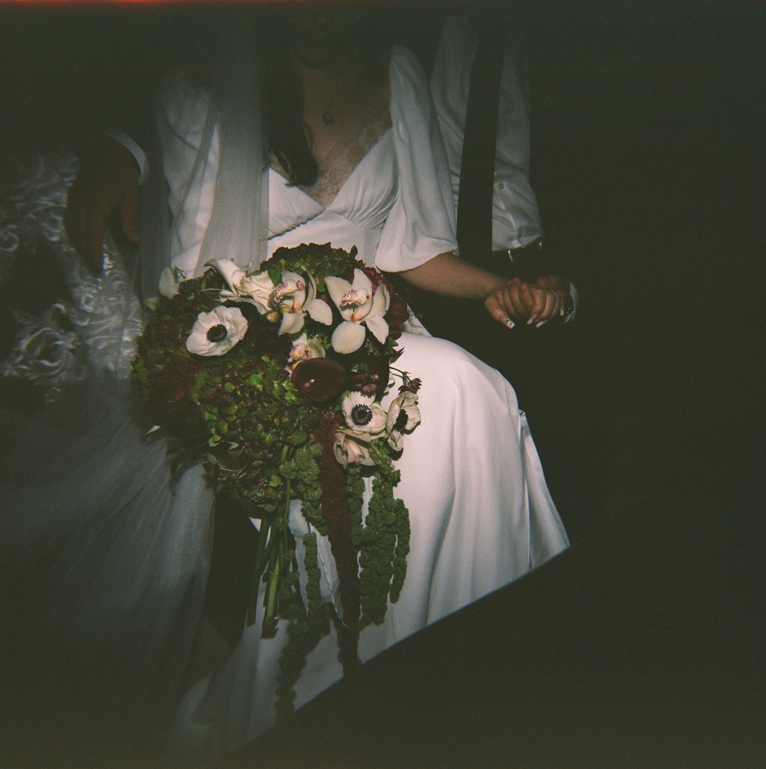 Bride in white wedding dress holding a lush bouquet of white and dark anemones with greenery at a dimly lit wedding.