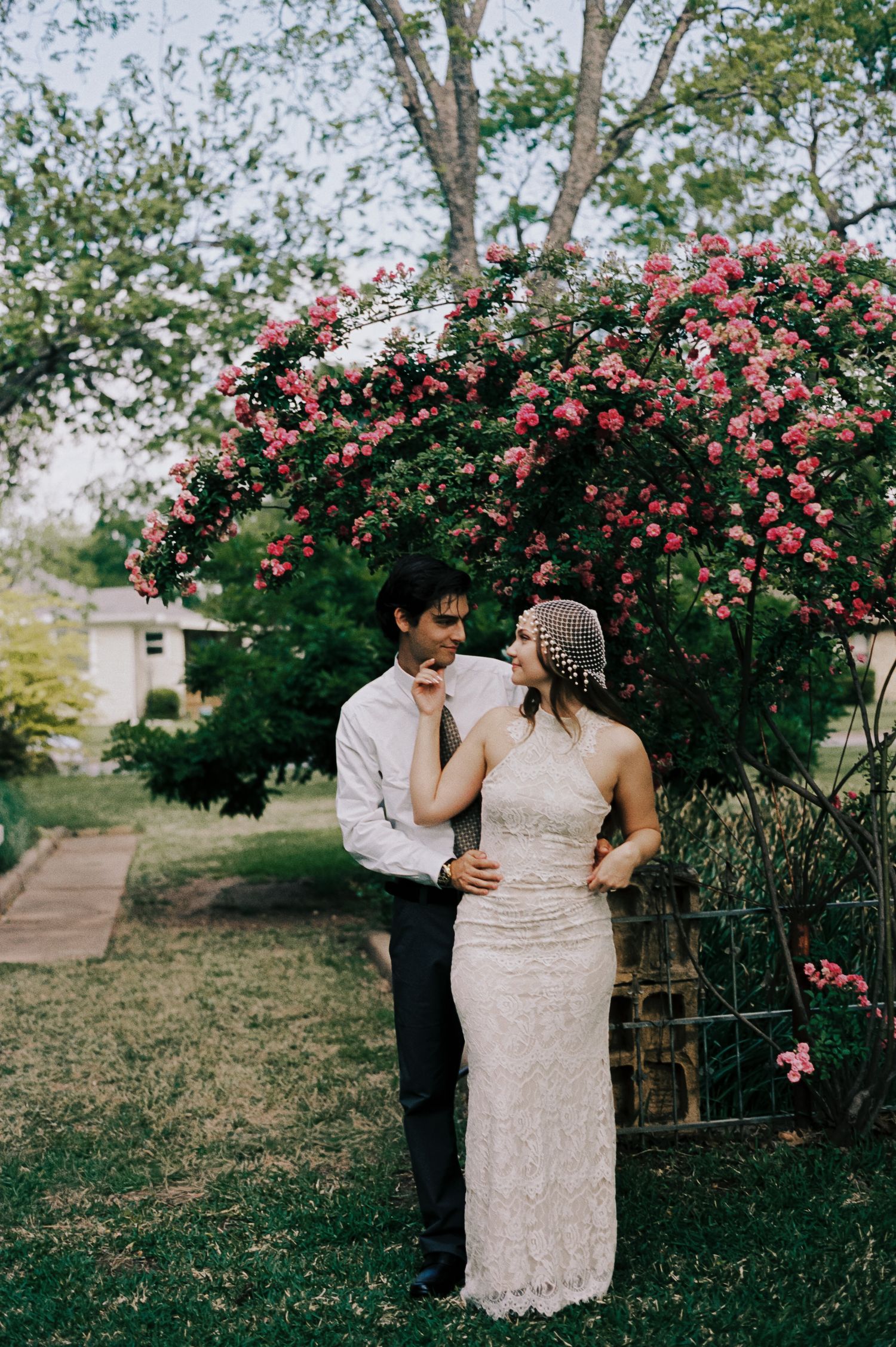 Couple in wedding attire sharing a romantic moment under a blooming pink rose arch in a lush garden.
