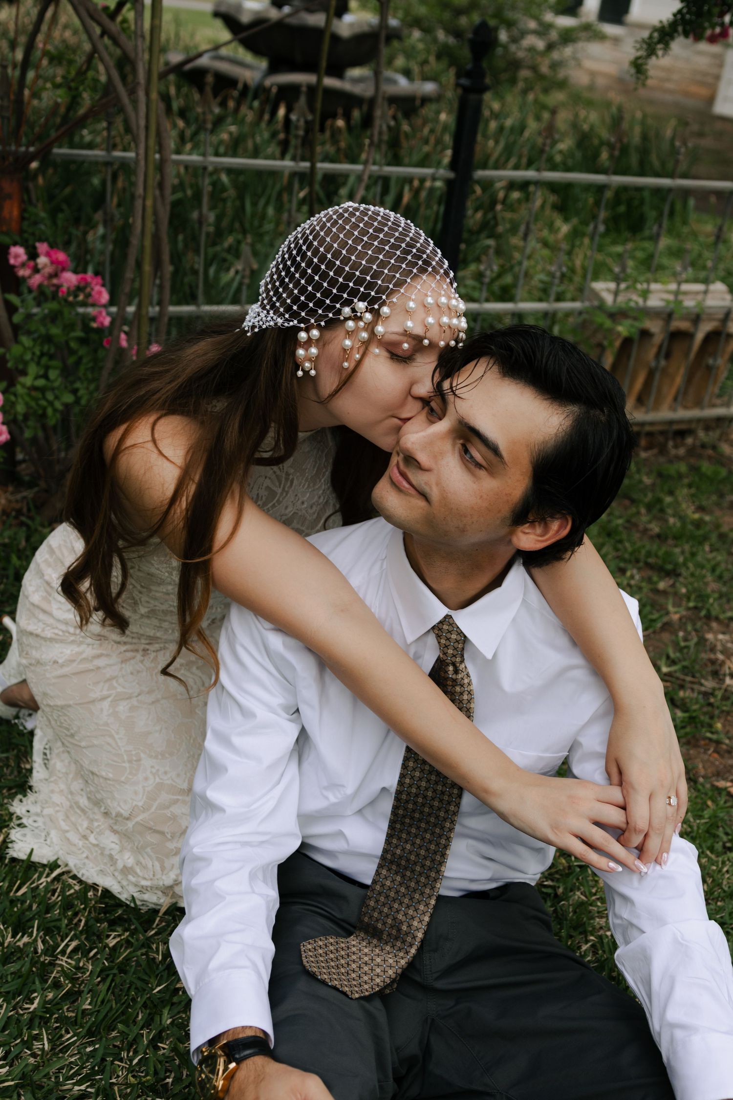 Bride in a pearl birdcage veil kisses groom on the cheek while sitting together on lush garden grass.