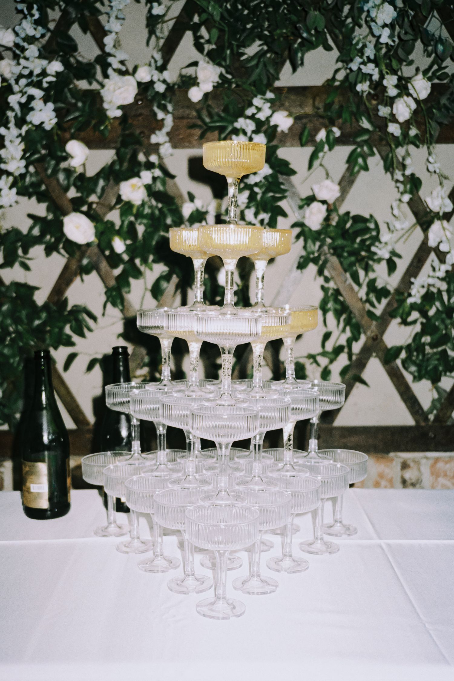 Elegant multi-tiered champagne tower with coupe glasses displayed before a floral greenery backdrop.