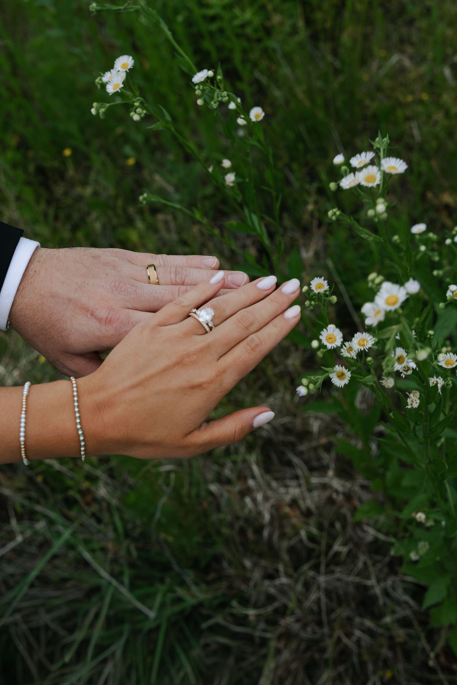 Newlywed couple holding hands showing wedding rings near white wildflowers in green grass.