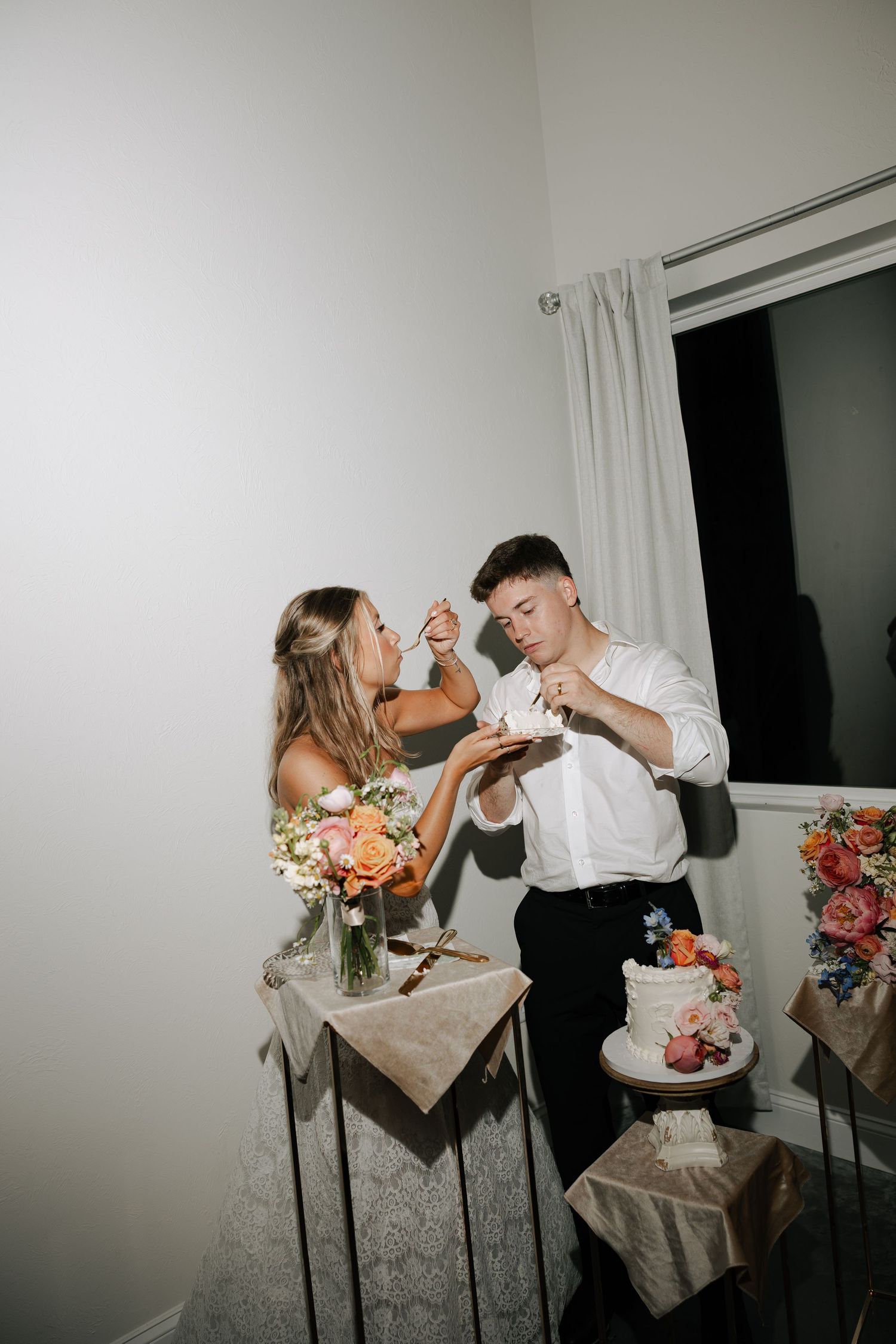 Bride and groom feed each other wedding cake at indoor reception beside floral decorated table.