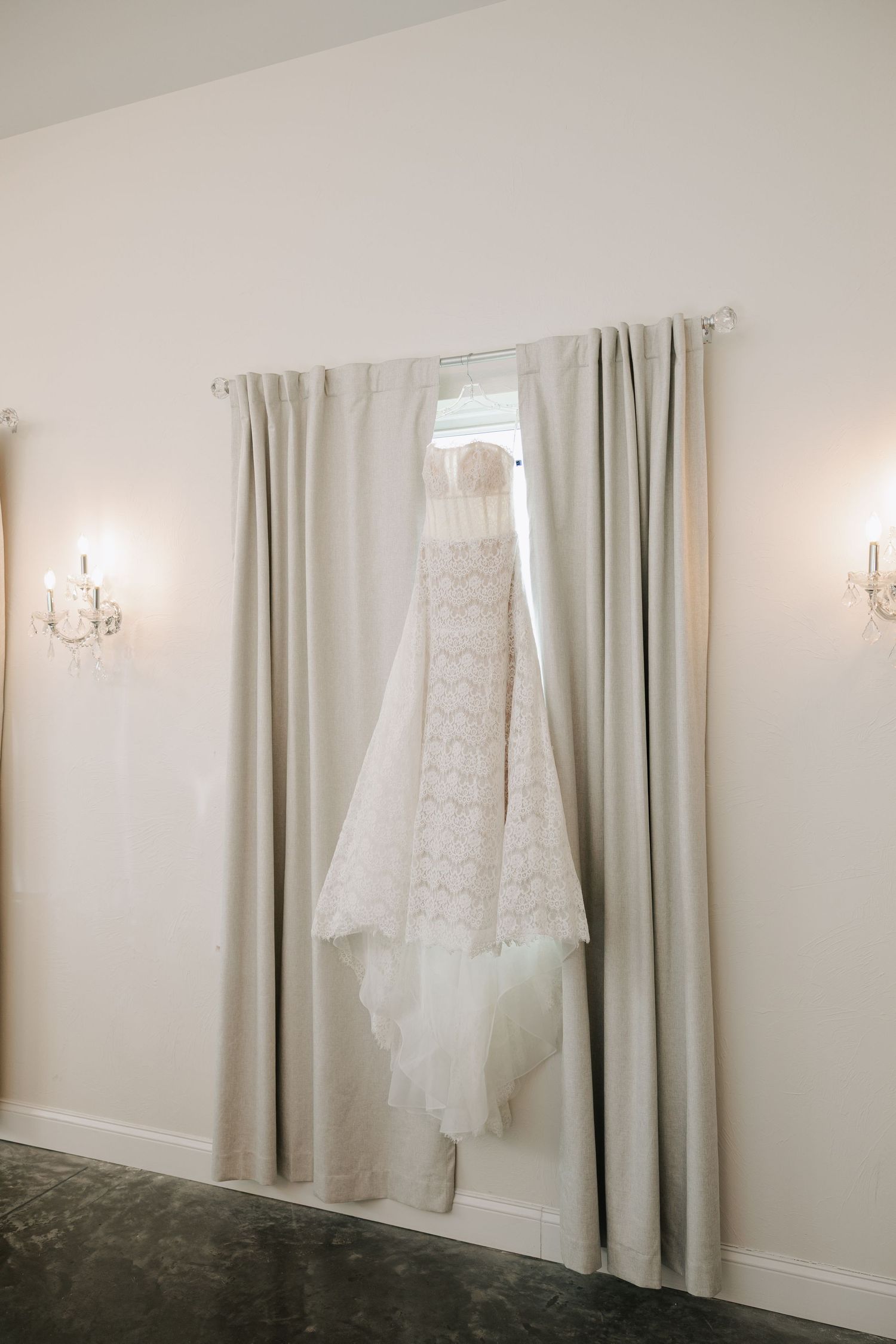Elegant lace wedding dress hanging in front of a window with gray curtains and crystal wall sconces.