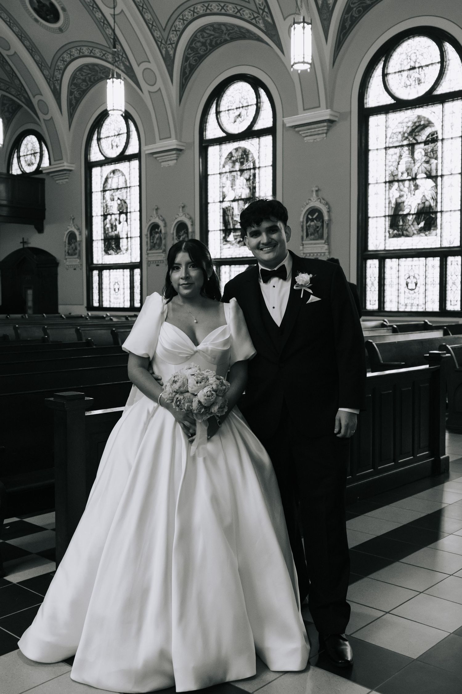 Black and white portrait of bride and groom smiling inside a stunning church with stained glass windows.