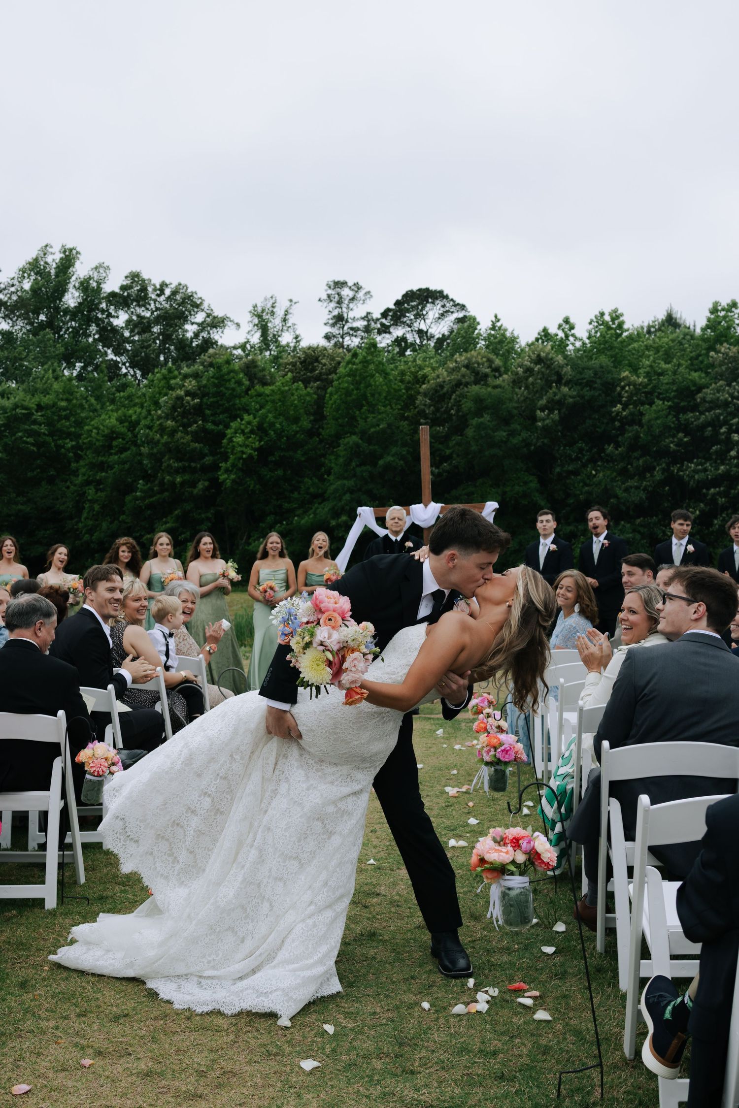 Groom dips bride for romantic kiss while guests cheer at outdoor wedding ceremony.