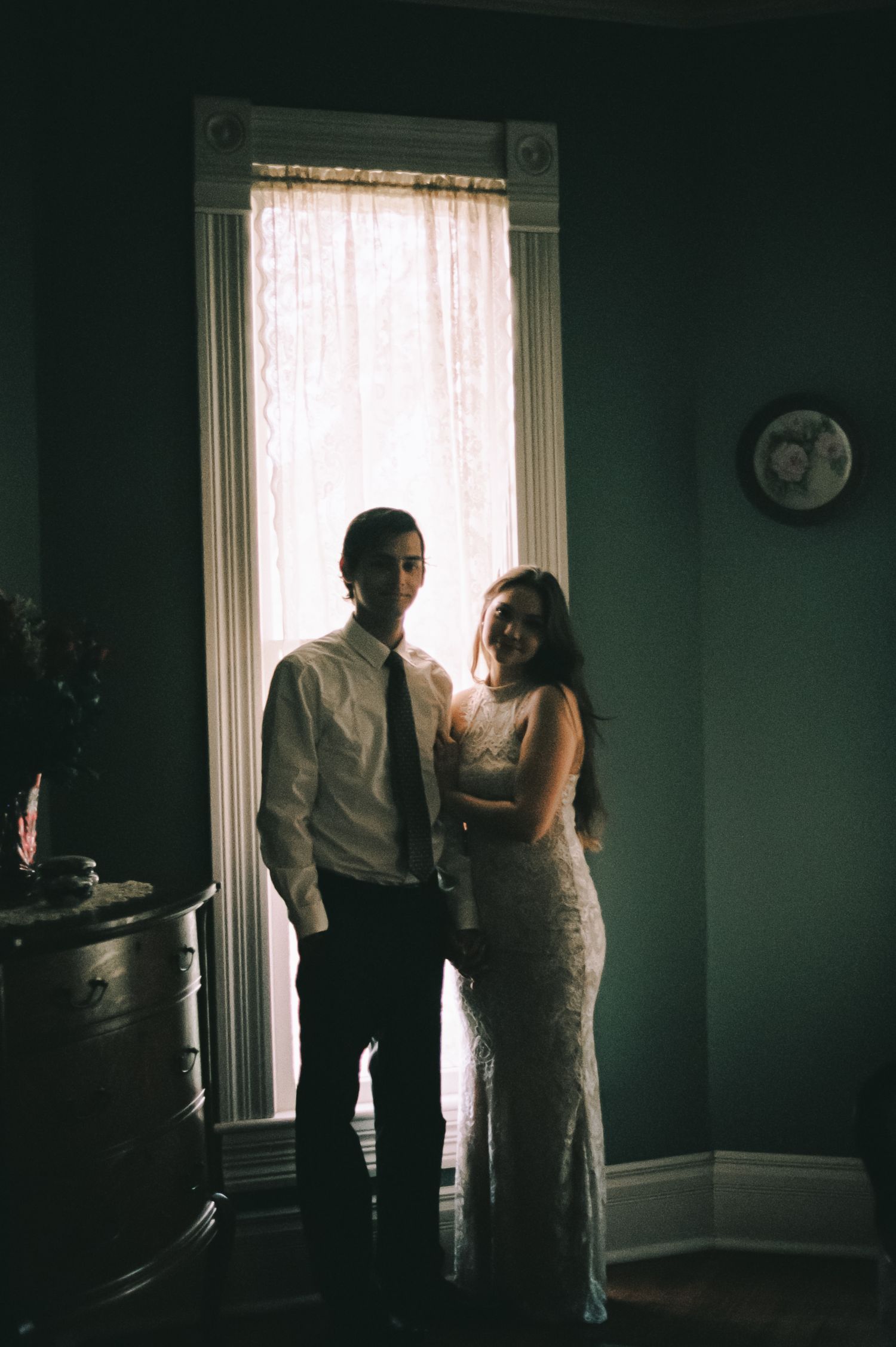 Couple in formal attire standing by a sunlit window in a dimly lit vintage room, elegant wedding portrait.