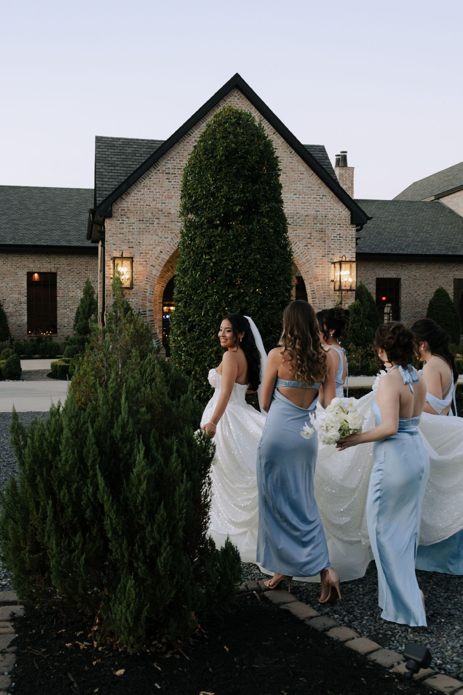 Bride in white gown walks with bridesmaids in blue dresses toward elegant brick wedding venue at dusk.
