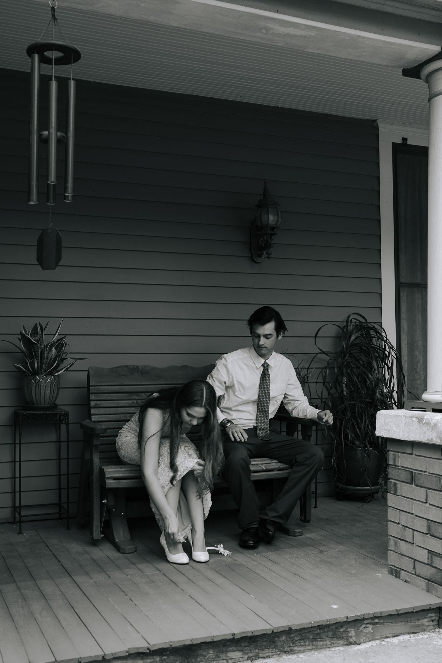 Black and white photo of a couple on a porch bench, woman tying her shoe while man in tie sits beside her.