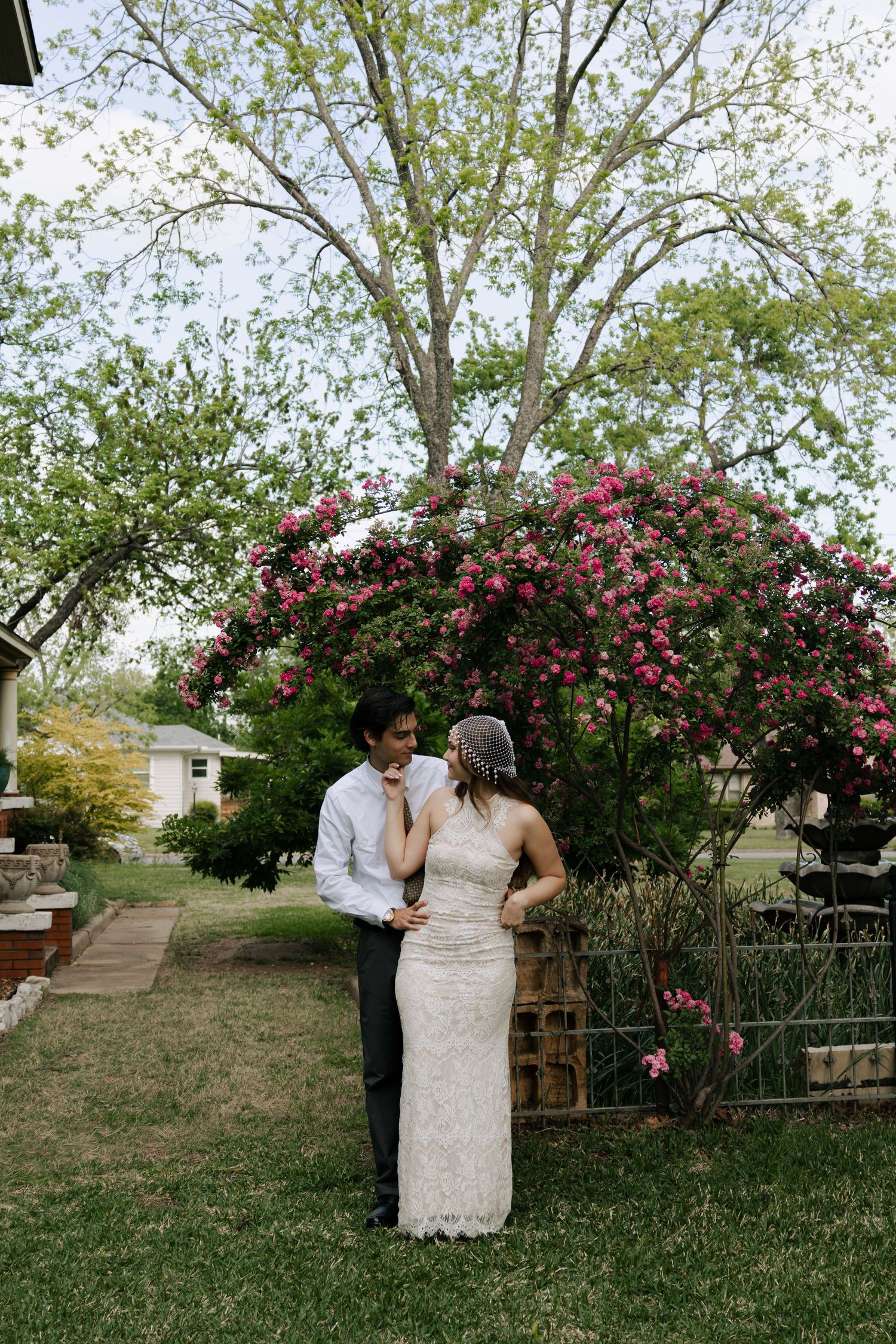 Bride and groom stand together in garden beneath blooming pink rose bush arch.