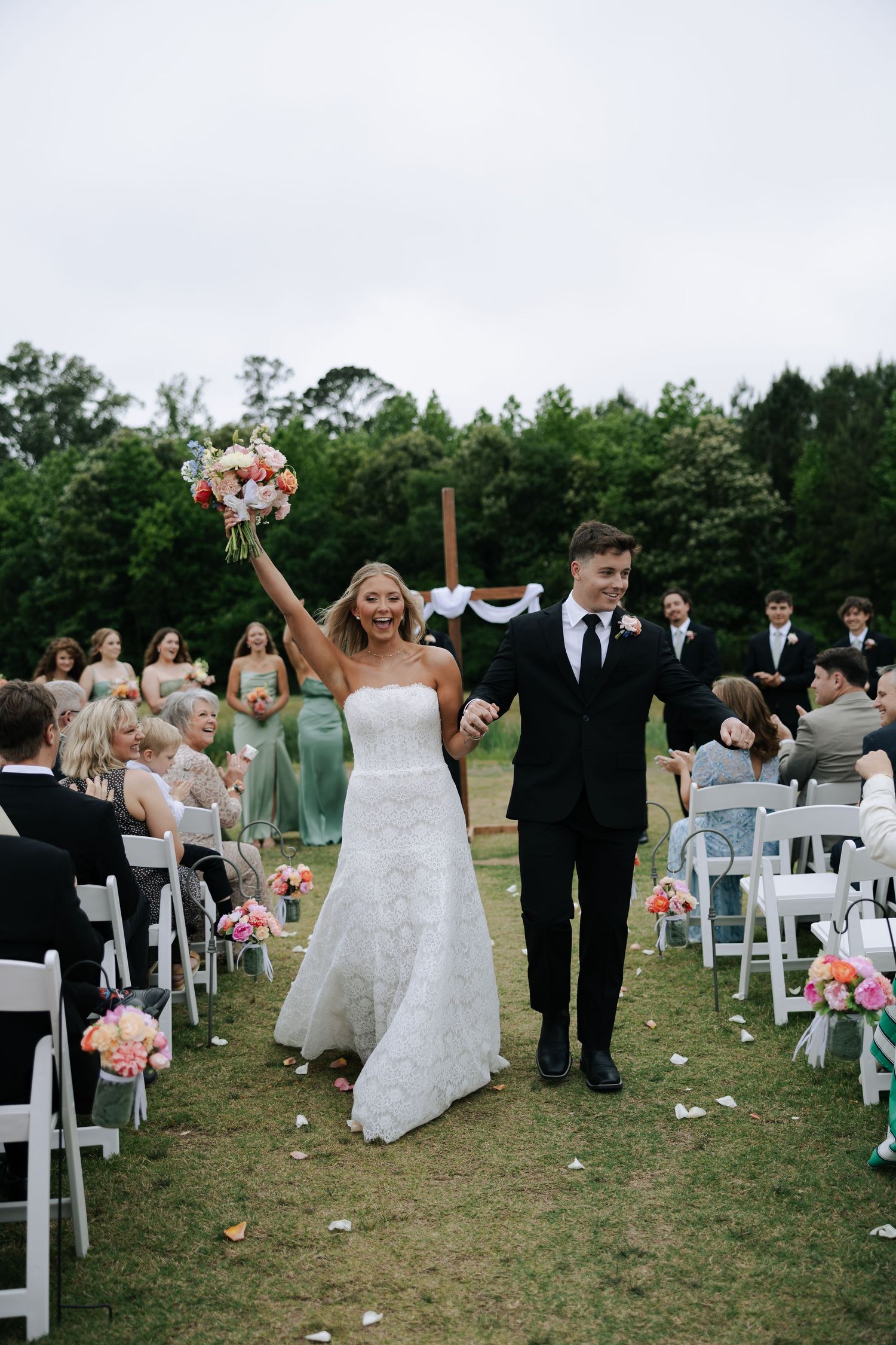 Joyful bride raising bouquet while walking back down the aisle with groom after outdoor ceremony.