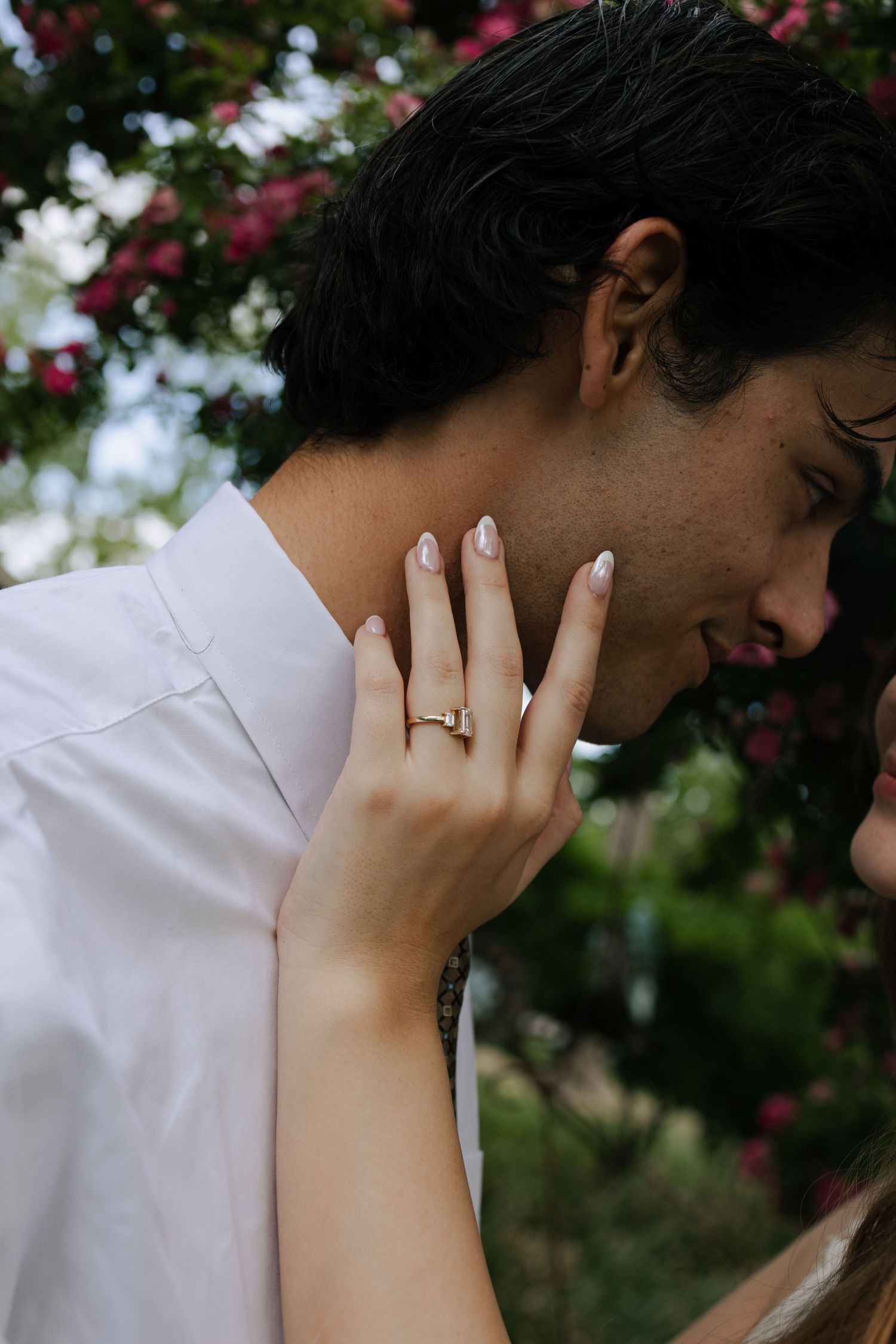 Couple embracing outdoors, woman displaying engagement ring with pink floral background.