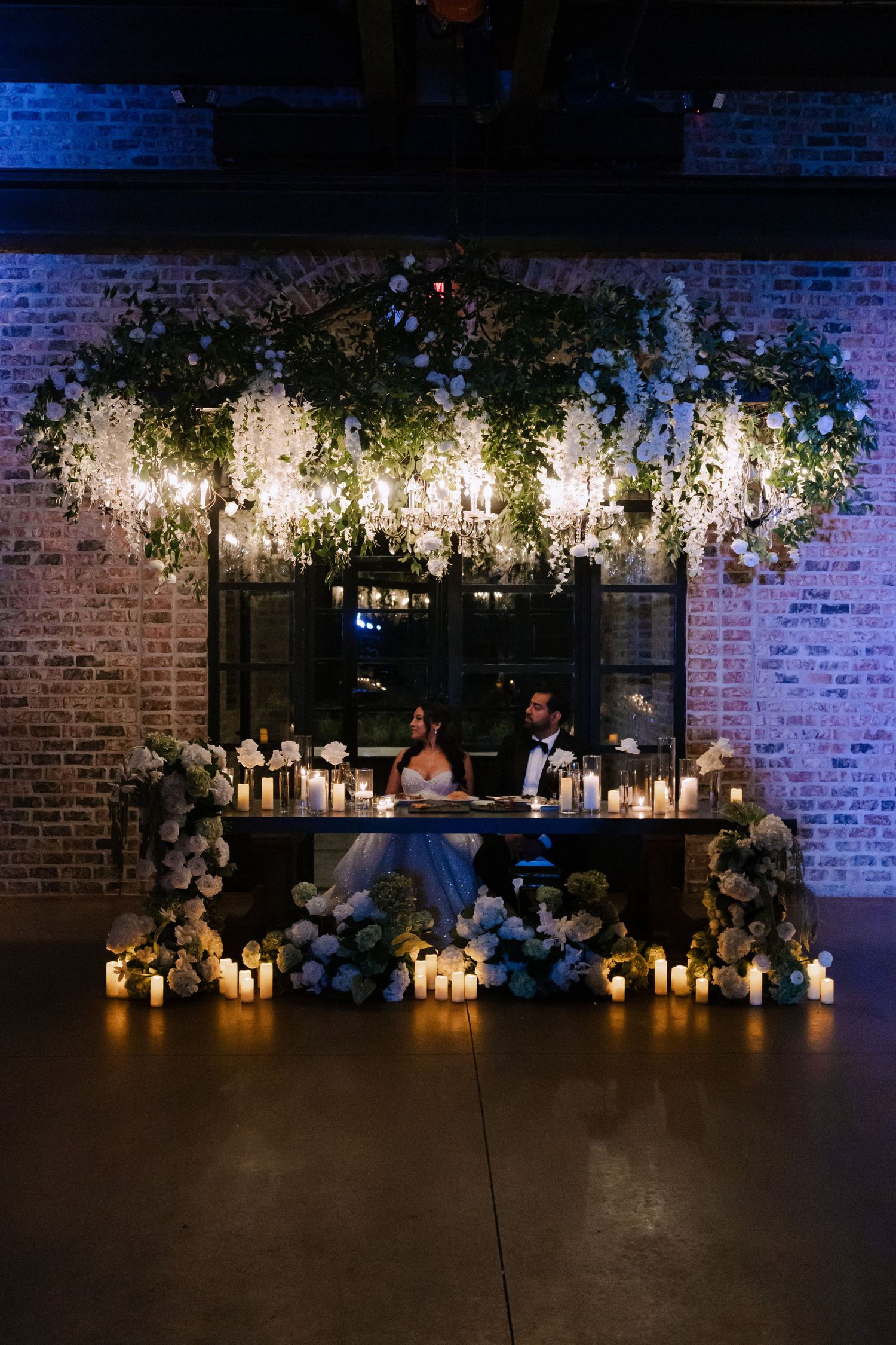 Bride and groom at candlelit sweetheart table adorned with hanging greenery and chandeliers in brick venue.