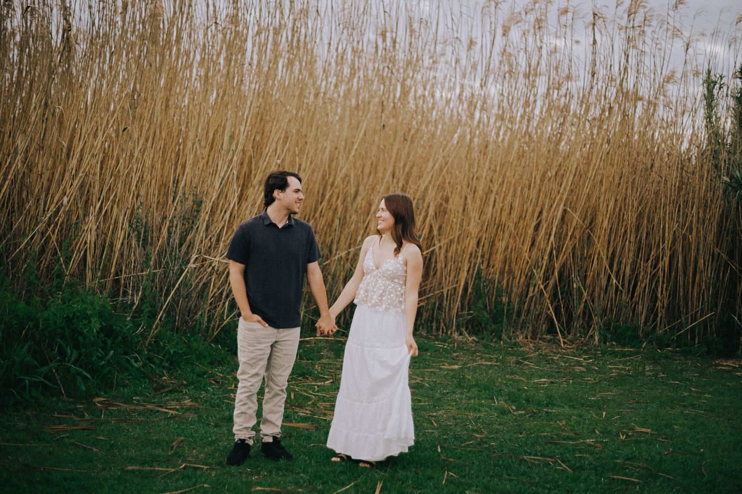 Engaged couple holding hands and smiling at each other in front of tall golden grass.