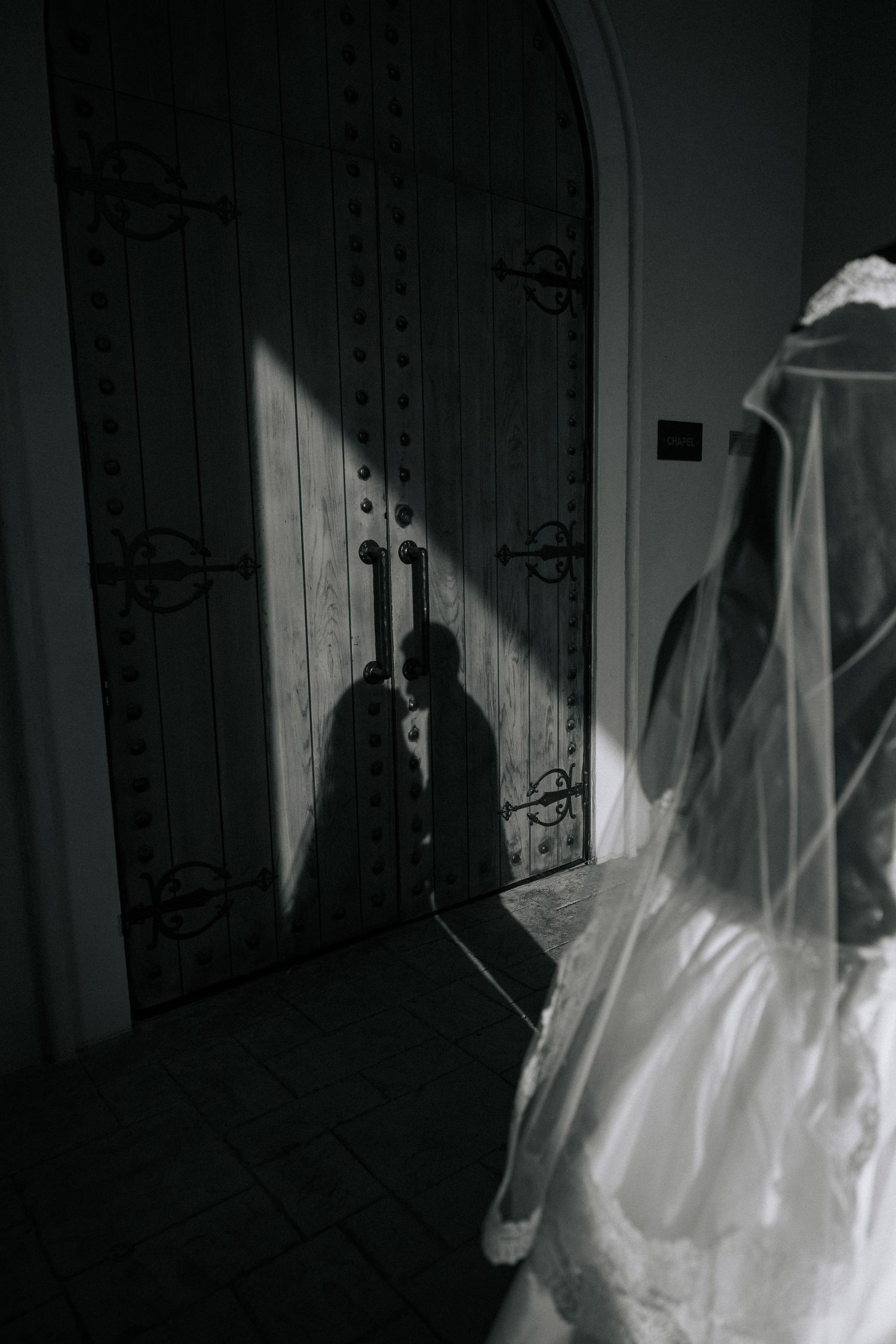 Black and white photo of bride in veil with silhouette of couple kissing cast on ornate wooden church doors.